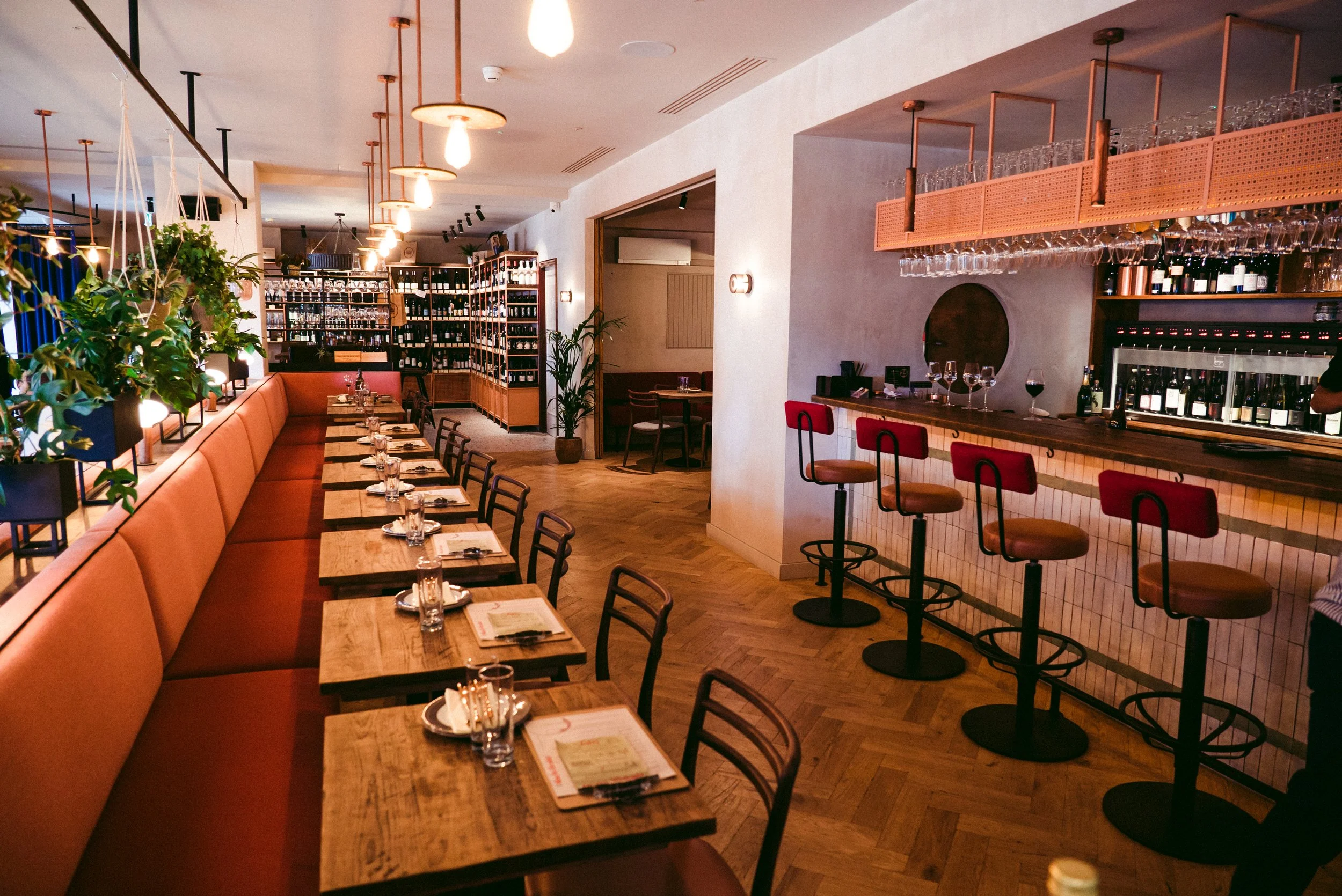 The bar and dining tables at Humble Grape Canary Wharf, wine shelving and red bar stools along the counter