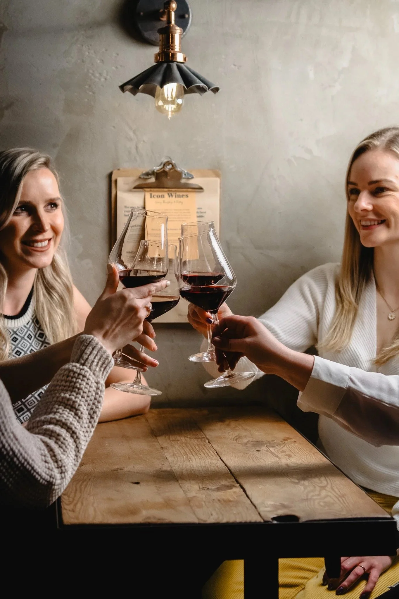 Two guests toast with glasses of red wine beneath a handwritten 'Icon Wines' notice at Humble Grape.