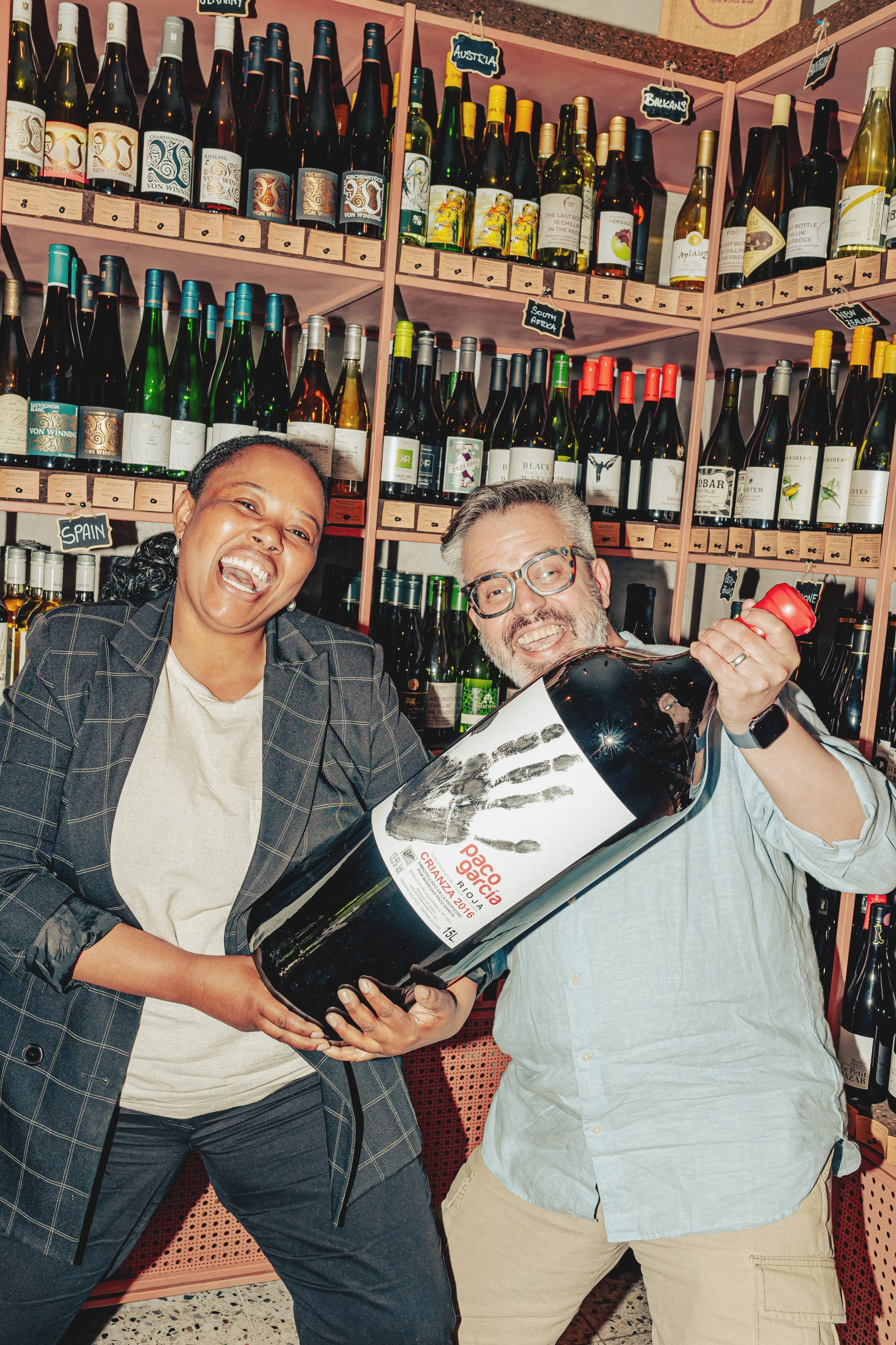 Two people, a woman and a man, smiling and holding a large bottle of red wine in a wine store with shelves filled with various bottles of wine behind them.