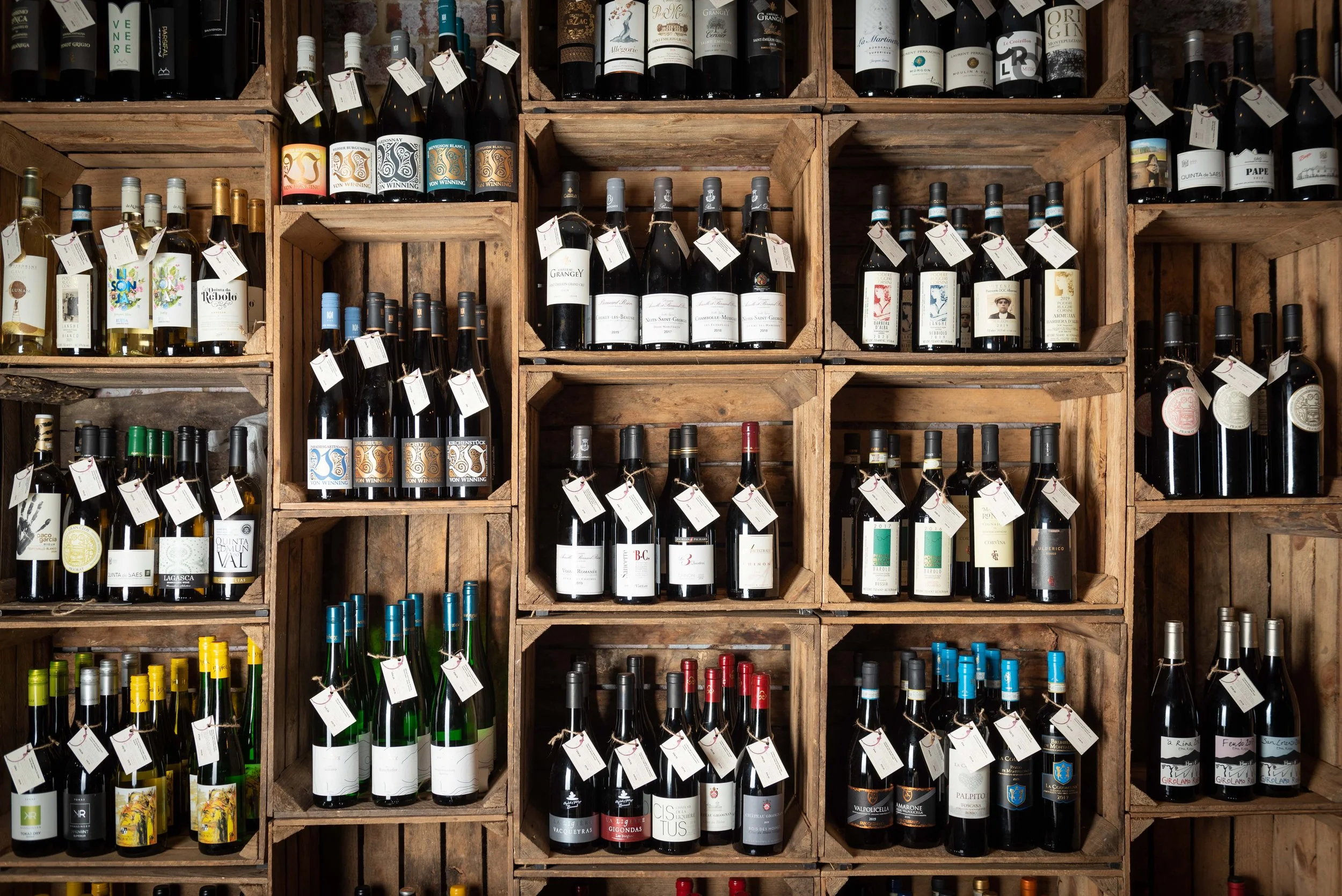 Wine bottles with handwritten price tags stacked in wooden crates on a Humble Grape shop wall.