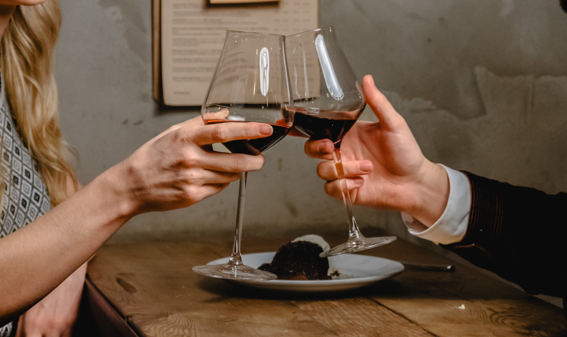 Two people clinking glasses of red wine at a dining table with a dessert plate in the background.