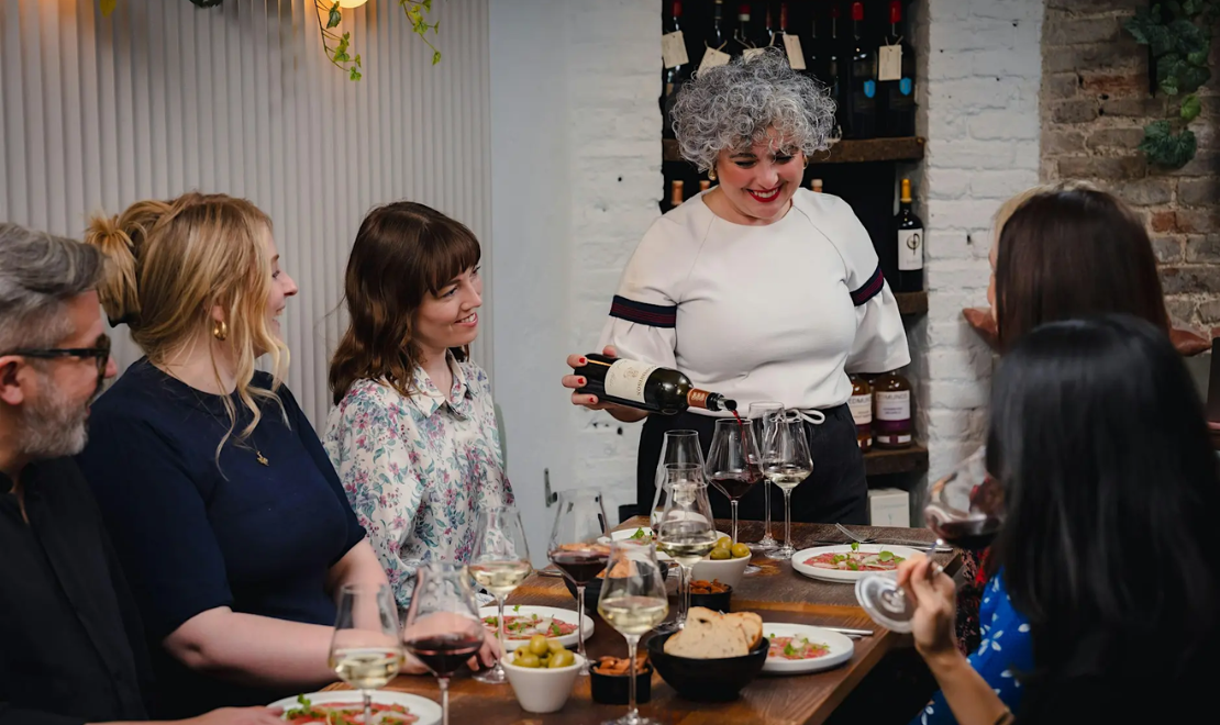 A group of people enjoying a wine tasting dinner with a female host pouring red wine, set in a cozy restaurant with wine bottles on the wall.