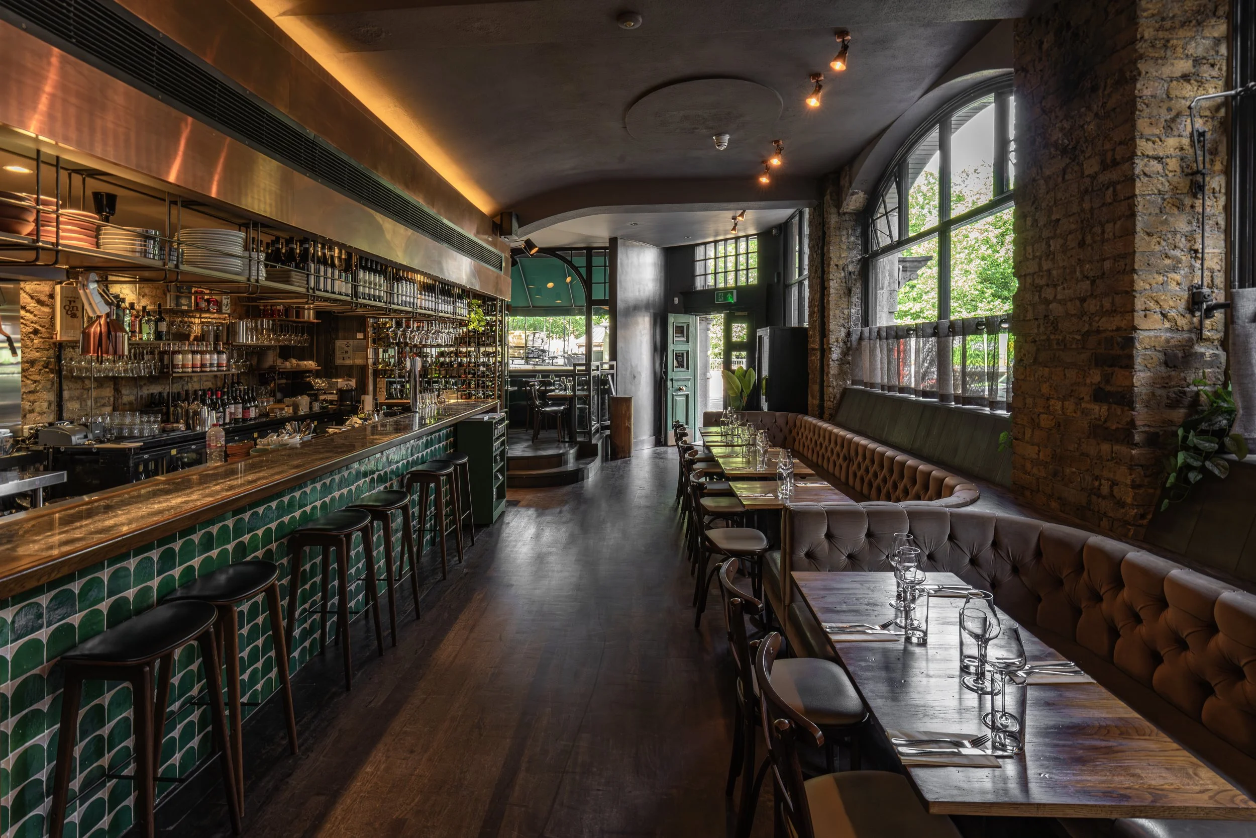 Interior of a modern restaurant with a bar counter on the left, featuring green and white patterned tiles, and a seating area with tables and chairs on the right, large windows with greenery outside, exposed brick walls, and warm lighting.