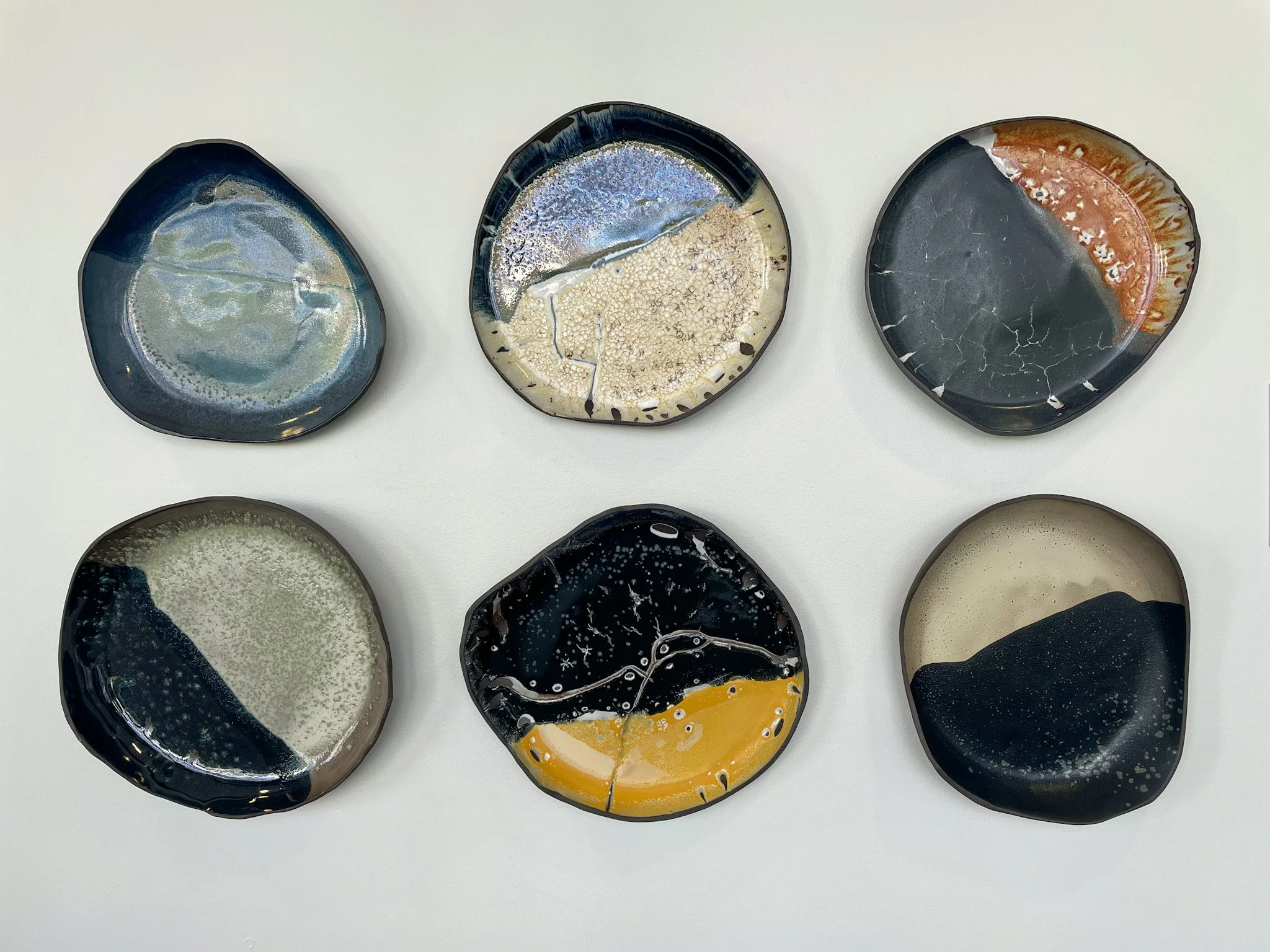 Six round, abstract ceramic plates with various glaze patterns, arranged in two rows against a white background.