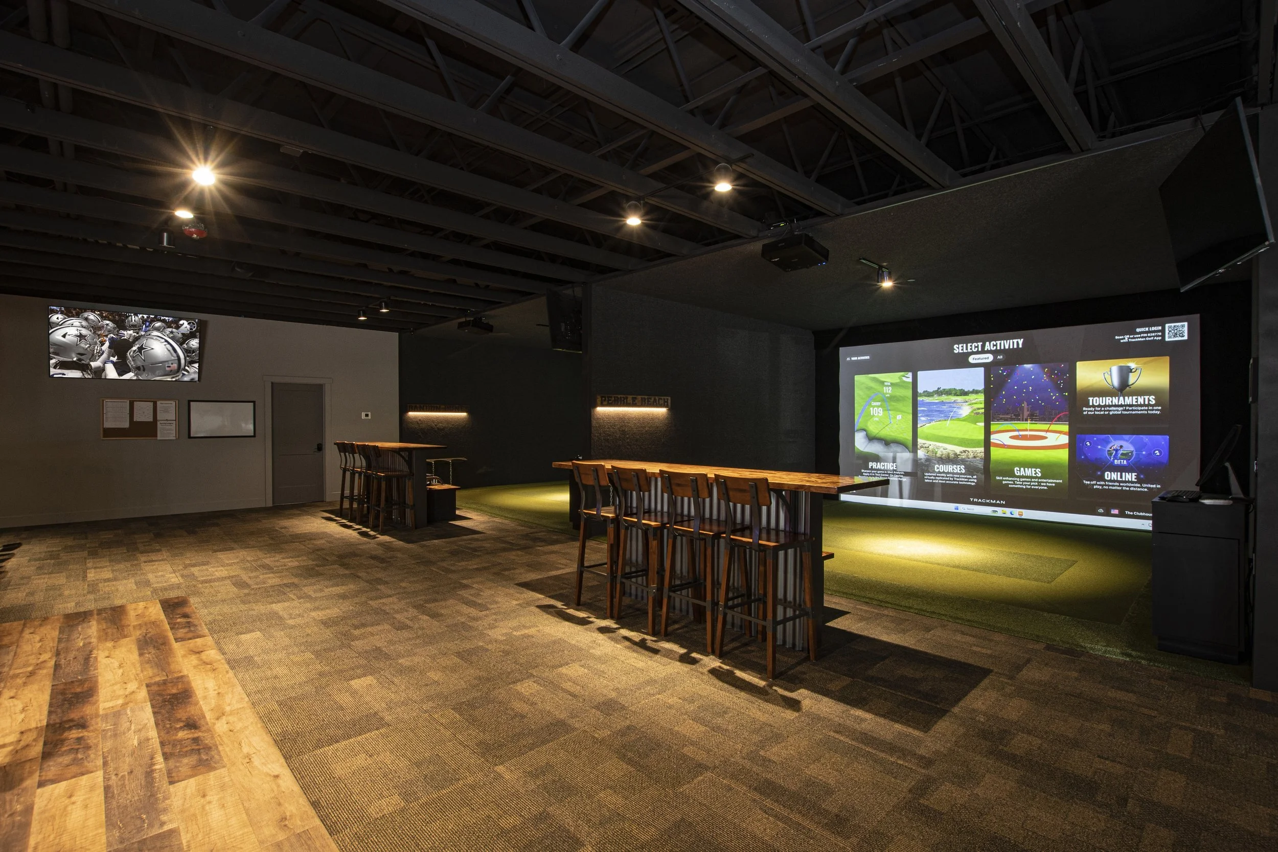 Indoor golf simulation room with chairs, a large screen displaying activity options, and a smaller wall-mounted screen showing helmets.