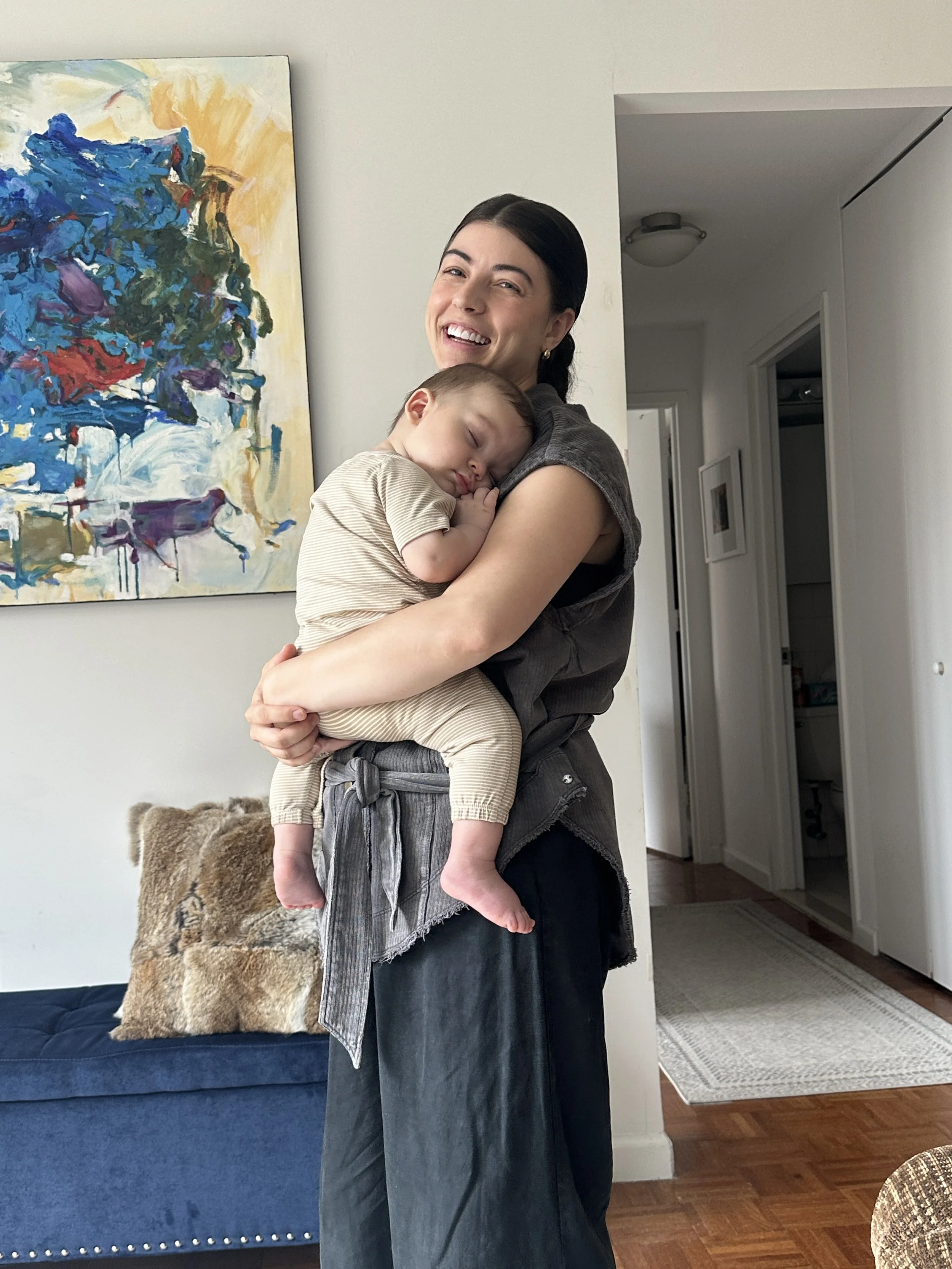 A woman with dark hair smiling and holding a sleeping young child in her arms, in a living room with artwork on the wall