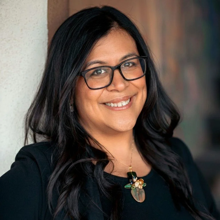 A woman with black hair, glasses, and a black top smiling, standing outdoors.