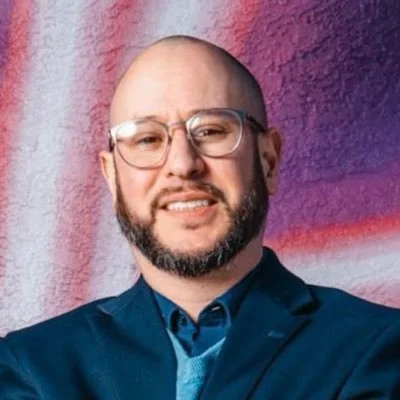 A man with a beard and glasses smiling in front of a colorful abstract background.
