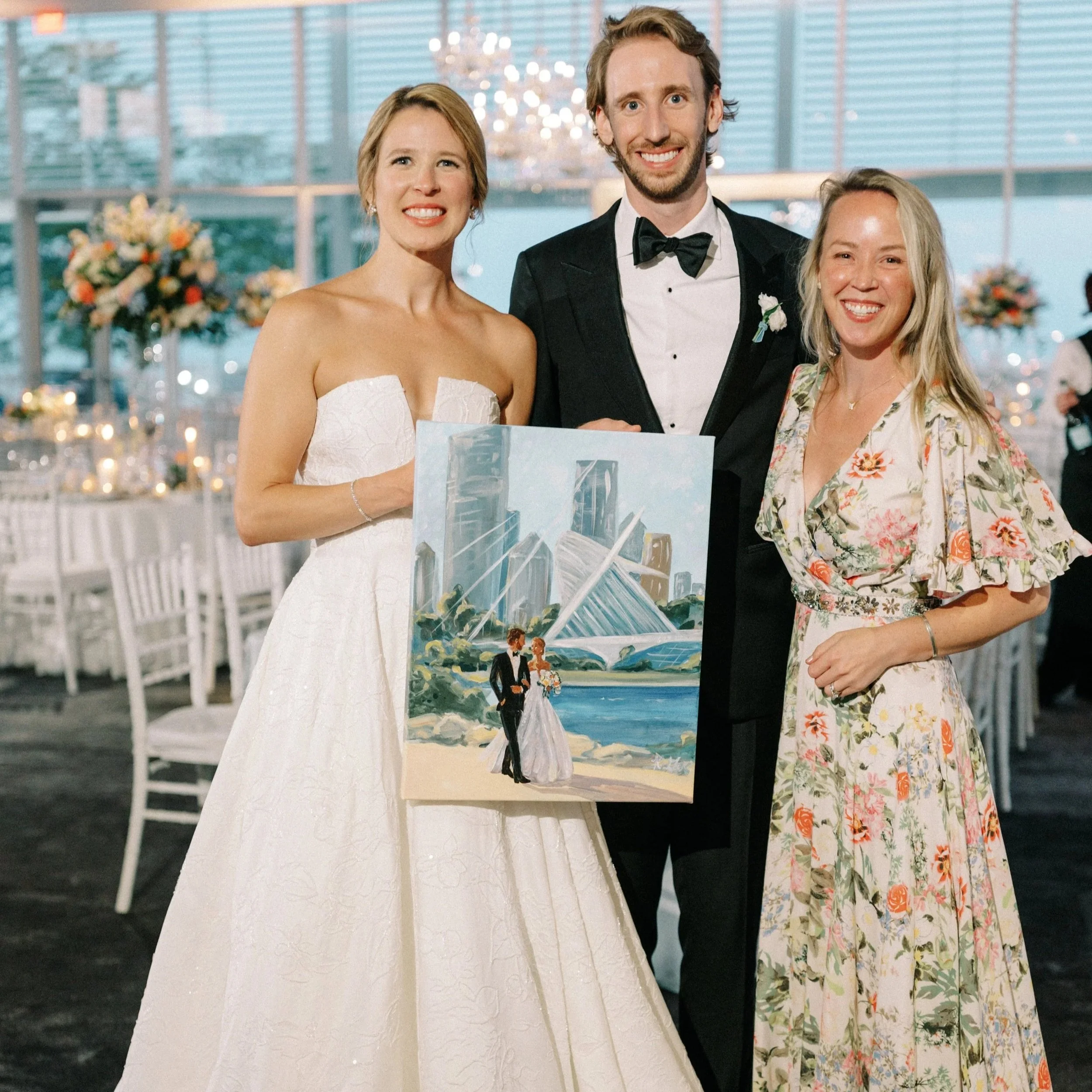 Bride and groom in wedding attire at night with city skyline in the background, holding a reflective, cityscape-themed painting.