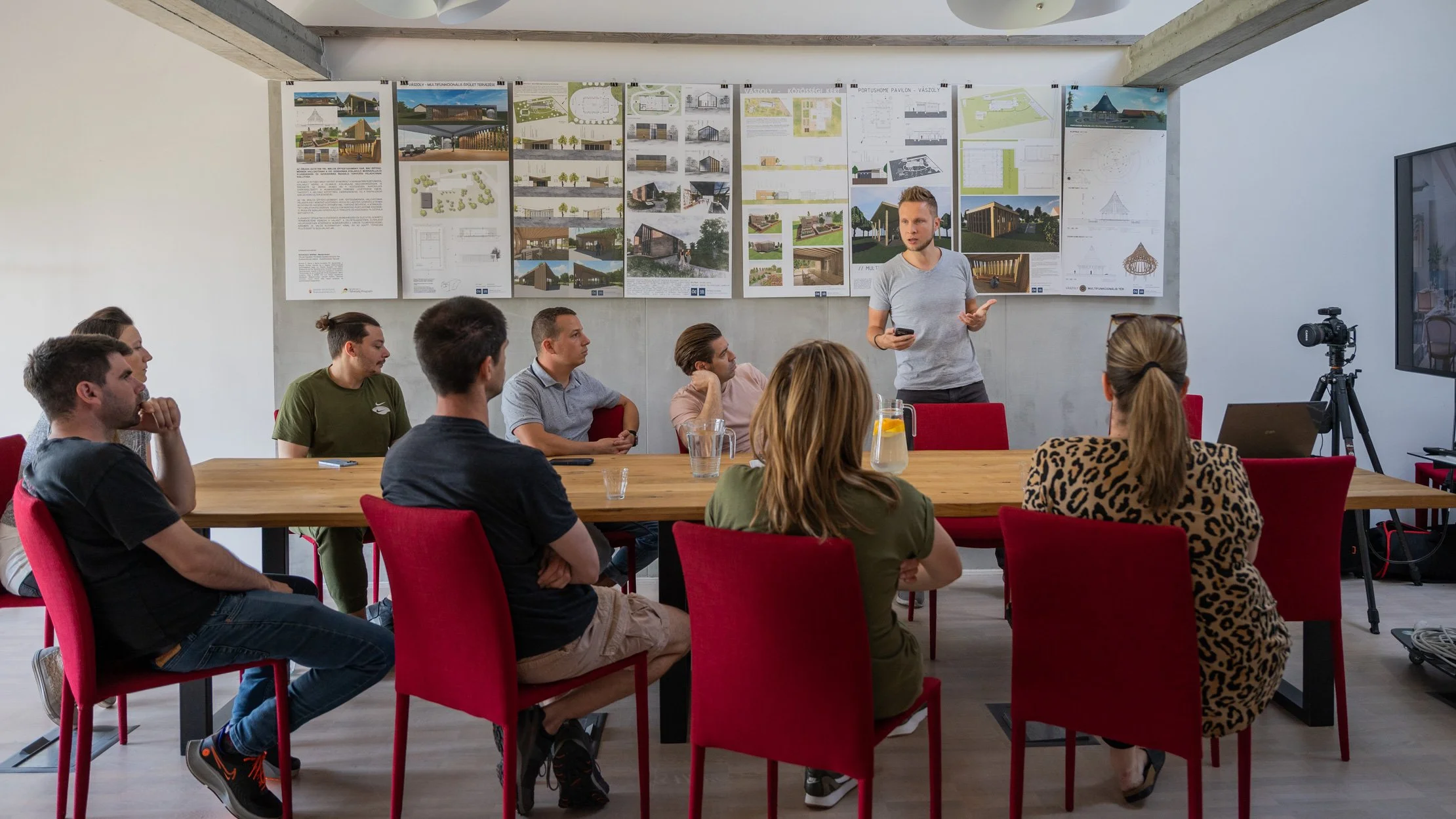 Ein Mann hält einen Vortrag vor einer Gruppe von Menschen in einem Konferenzraum, mitarchitektonischen Plänen und Entwürfen an der Wand