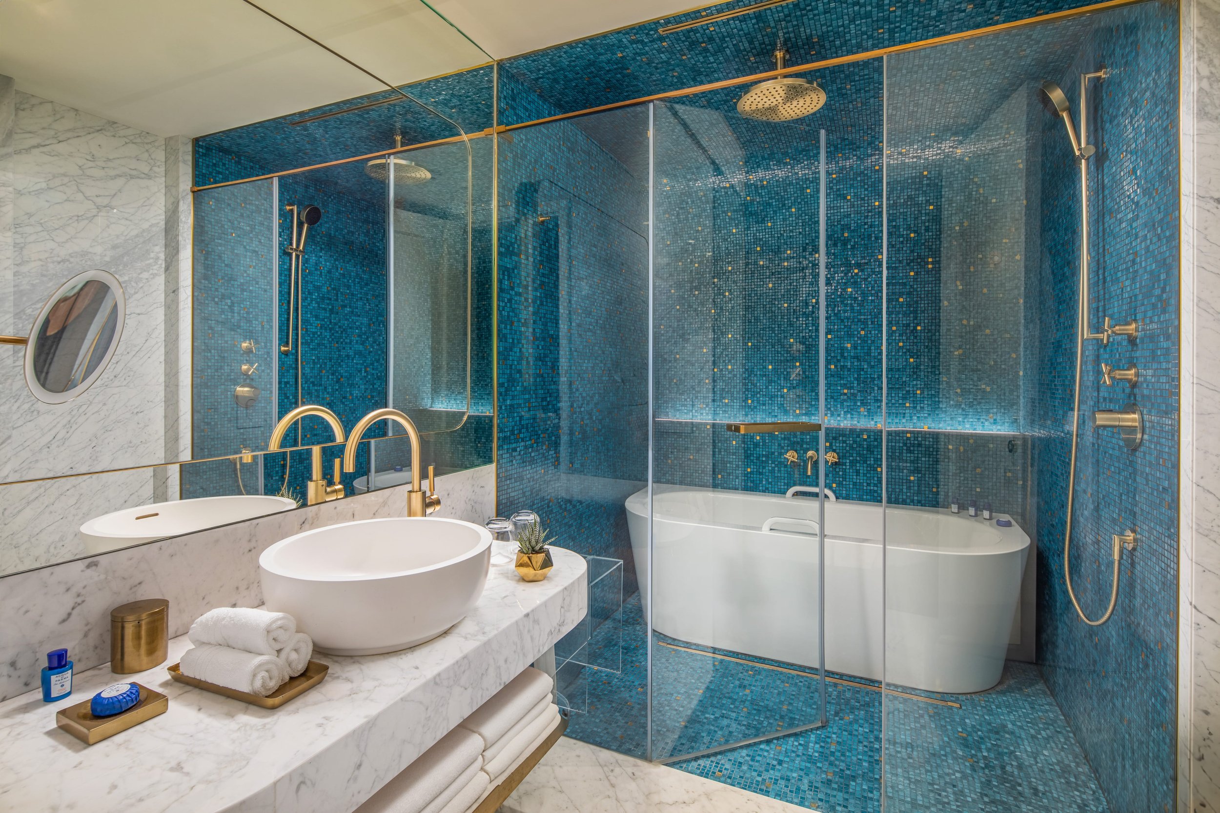 Moderner Badezimmer mit blauer Mosaikwand, freistehende Badewanne, große Spiegelwand, Waschtisch mit weißer Schale, goldene Armaturen, kleine Pflanzen, Handtücher und Badeutensilien.