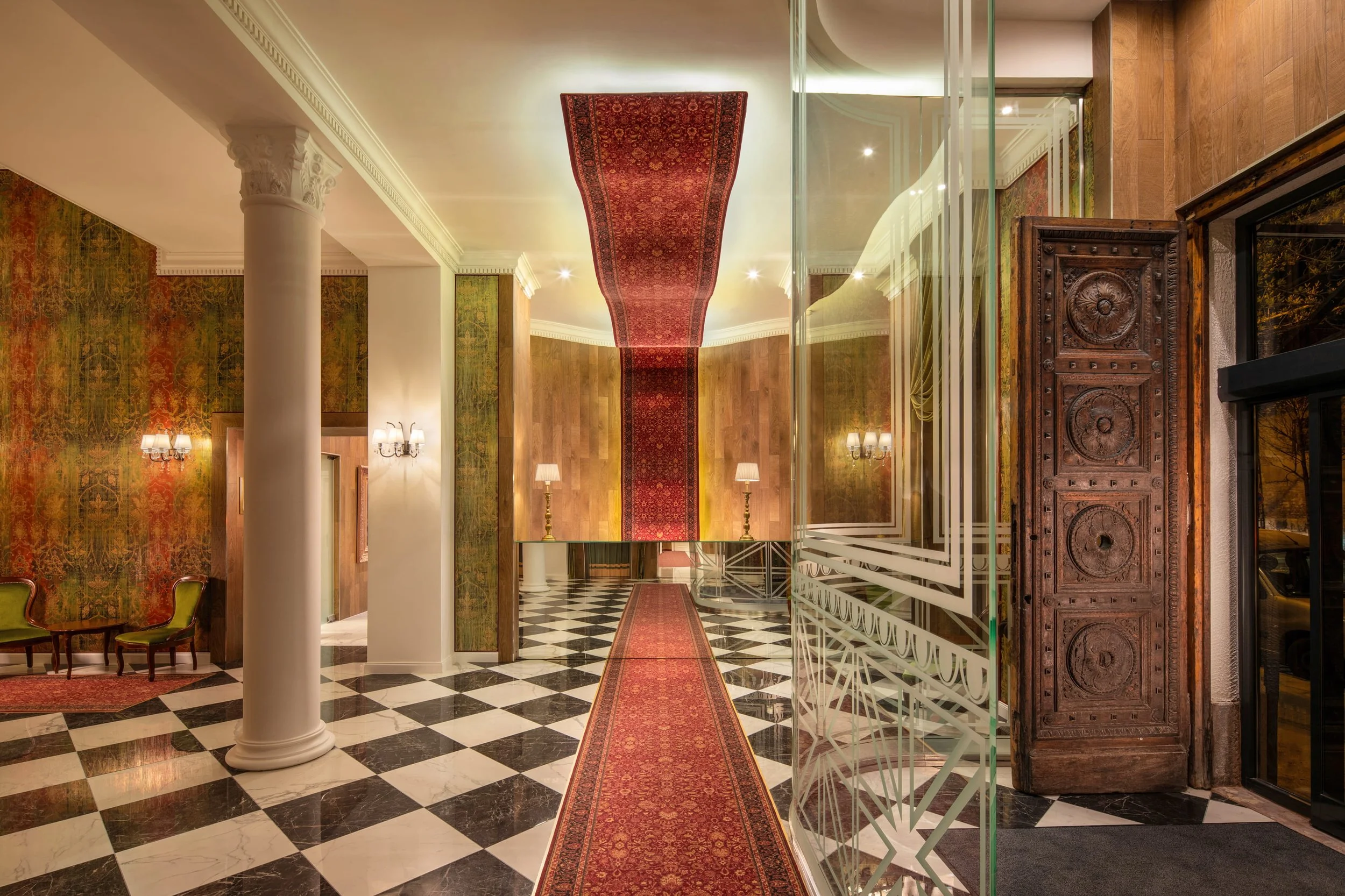 Elegante Hotelhalle mit schwarzen und weißen Fliesen, rotem Läufer, goldenen Stehlampen, grünen Stühlen, und einer antiken Holztür
