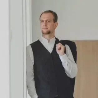 Ein Mann steht an einer Tür, trägt einen weißen Hemd und eine schwarze Weste, hält eine Jacke über die Schulter und blickt ernst in die Kamera.