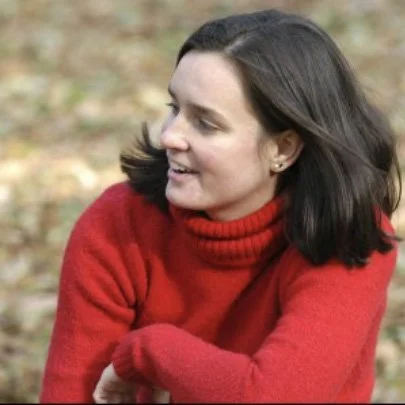 Frau mit dunklen Haaren, rotem Pullover, lachend im Freien mit Herbstlaub im Hintergrund.