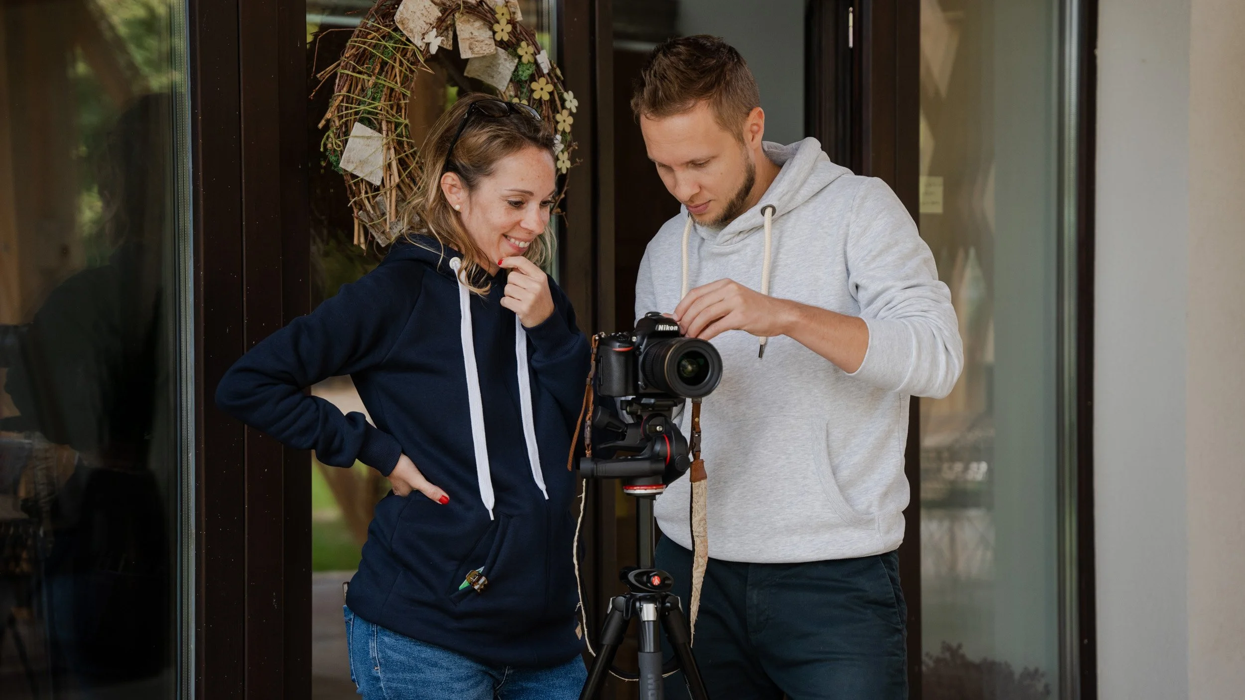 Zwei junge Menschen schauen auf eine Kamera auf einem Stativ, während sie gemeinsam vor einer Tür stehen.
