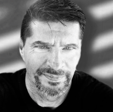 Ein Mann mit dunklem Haar und Bart, lachend, im Schwarz-Weiß-Foto aufgenommen.