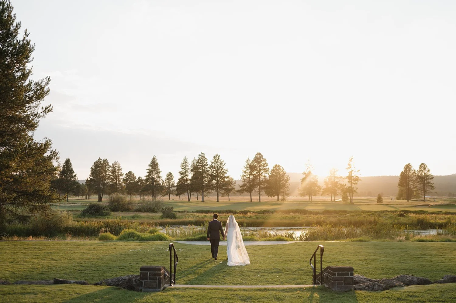 A bride and groom walking hand in hand on a grassy area towards a pond with a scenic landscape of trees and hills during sunset.