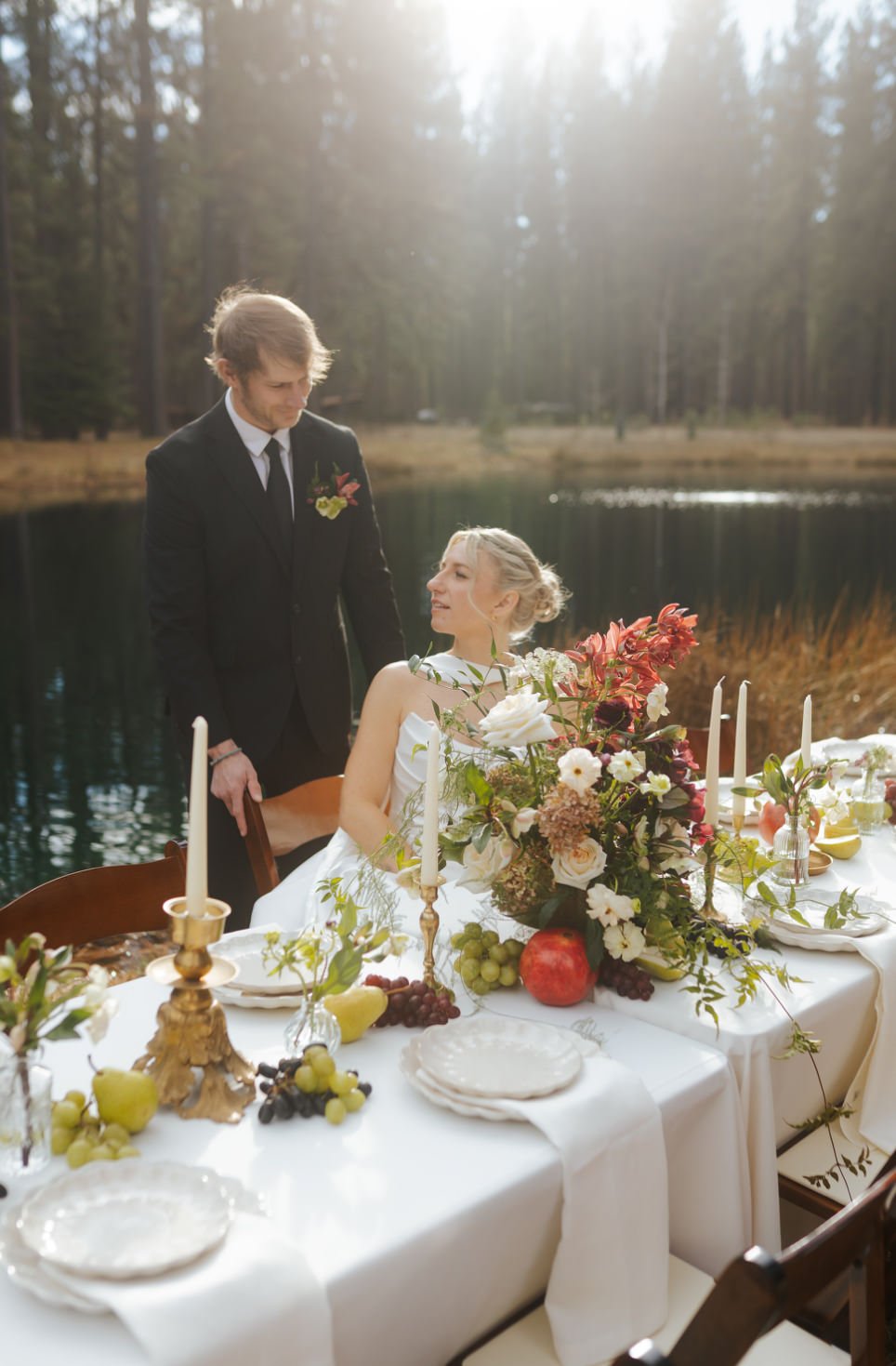 A couple dressed in wedding attire at an outdoor reception table near a lake, with trees in the background and decorated with candles, flowers, and fruit.