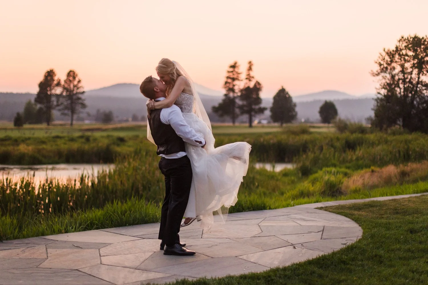 A couple dancing outdoors on a stone path during sunset, with a lake and trees in the background.