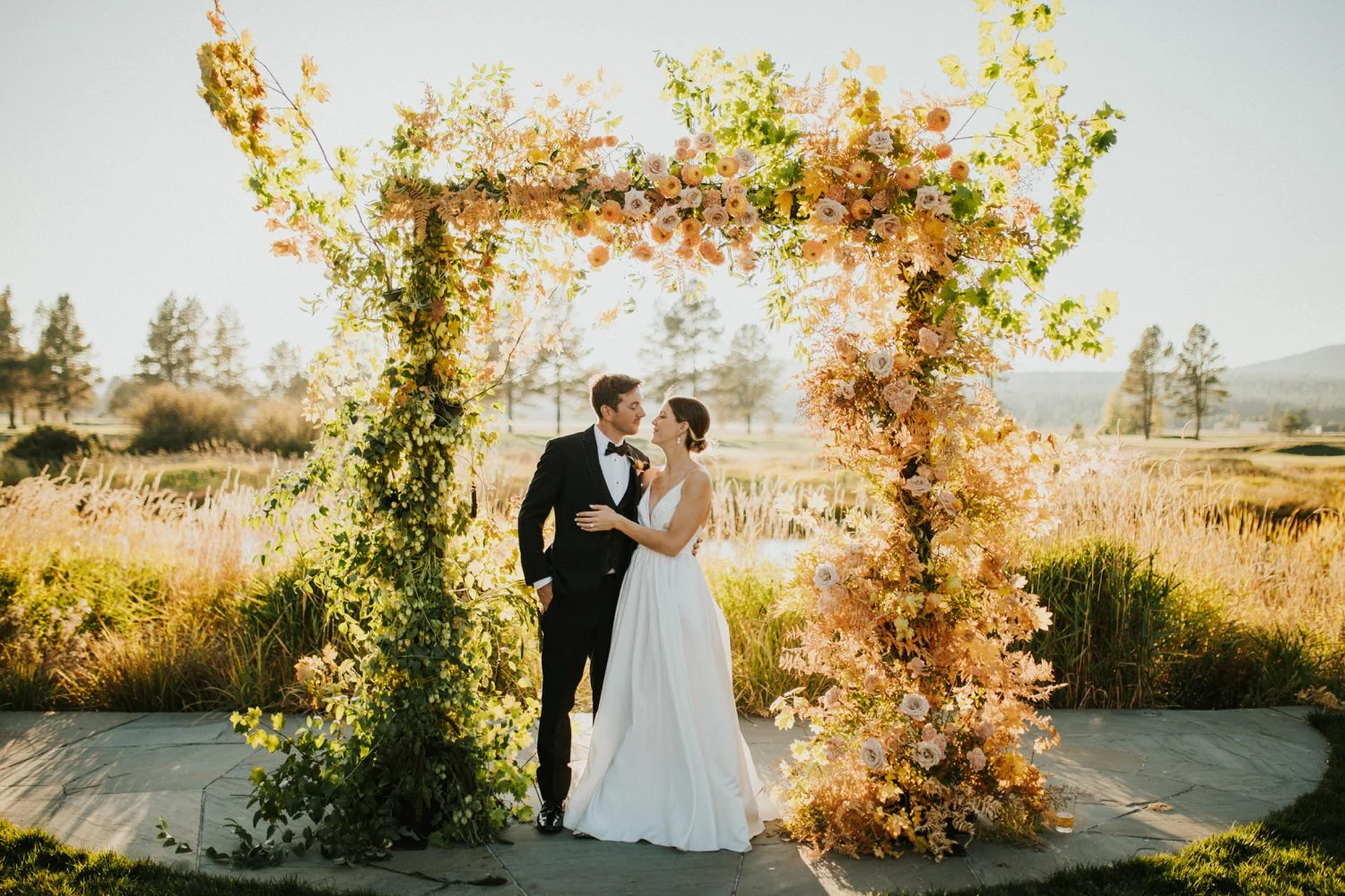A bride and groom standing under a floral wedding arch outdoors during sunset, surrounded by greenery and trees in the distance.