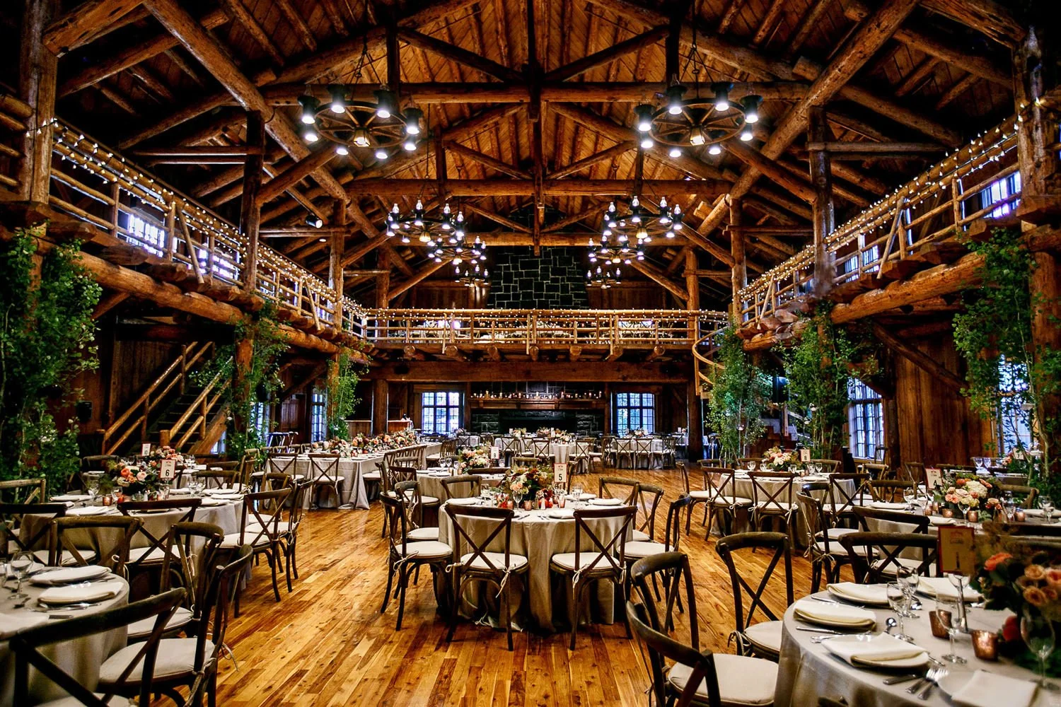 A rustic, wooden banquet hall decorated for an event with round tables, floral centerpieces, and string lights.