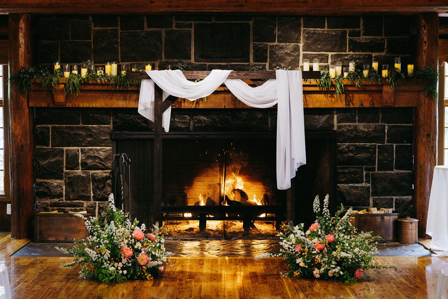 A decorated fireplace mantle with candles, greenery, and white fabric draped over dark wood beams, with floral arrangements in front on a wooden floor.