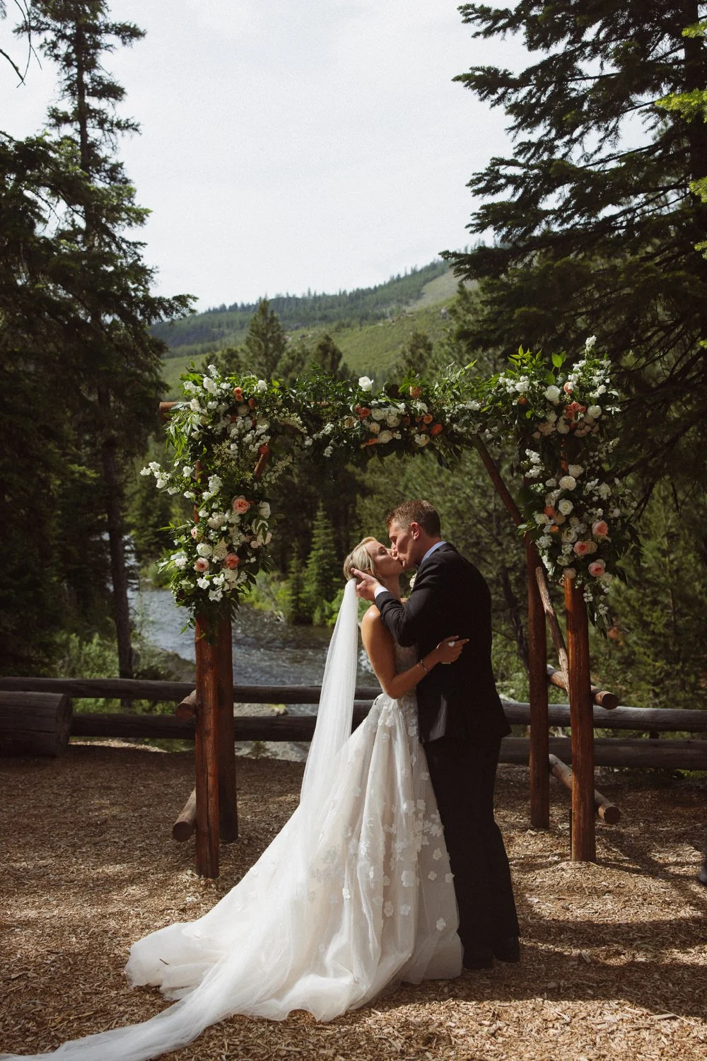 Bride and groom share a kiss under a floral wedding arch outdoors surrounded by trees and mountains.