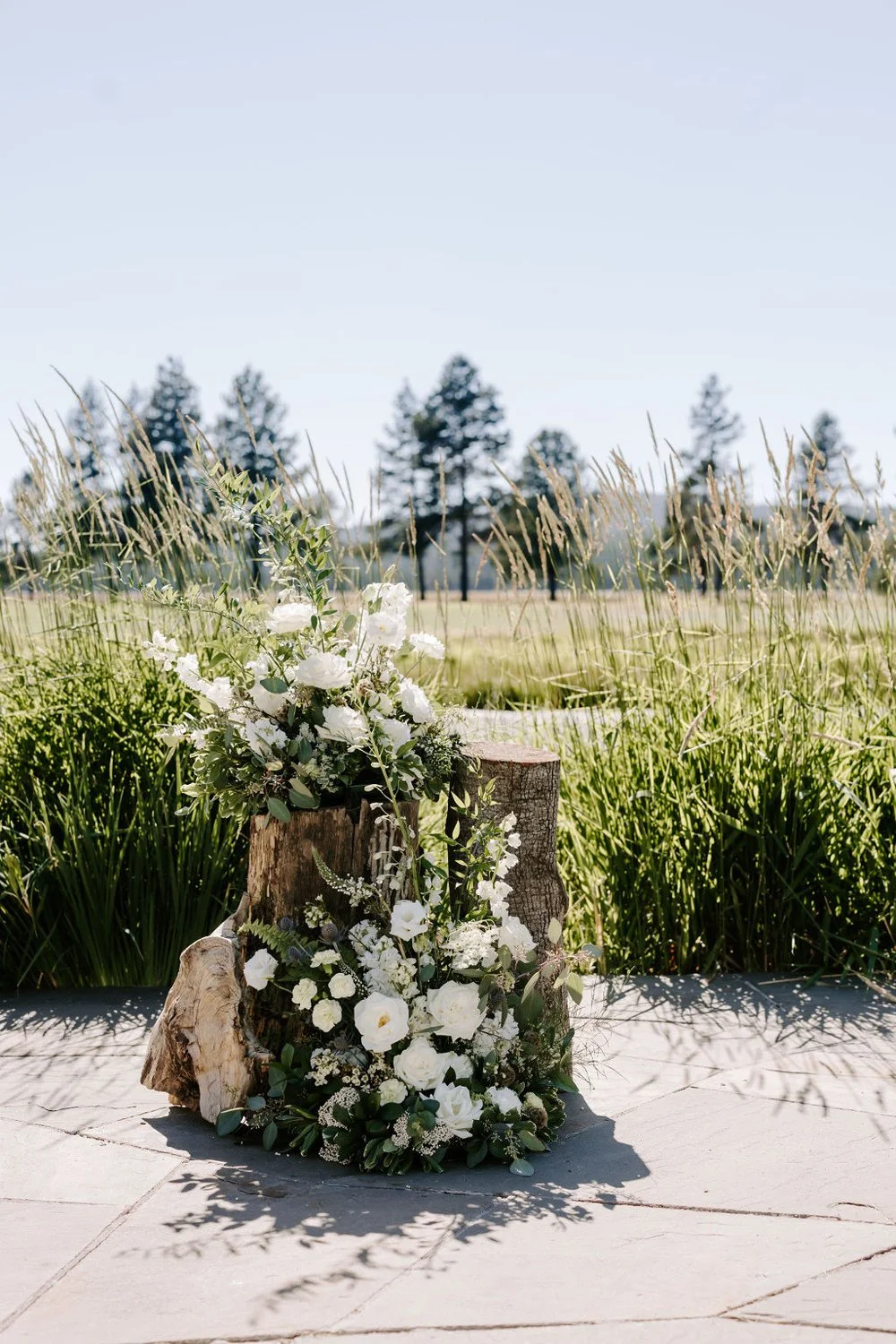 a floral arrangement with white flowers and greenery on a wooden stump outdoors, with tall grass and trees in the background under a clear blue sky
