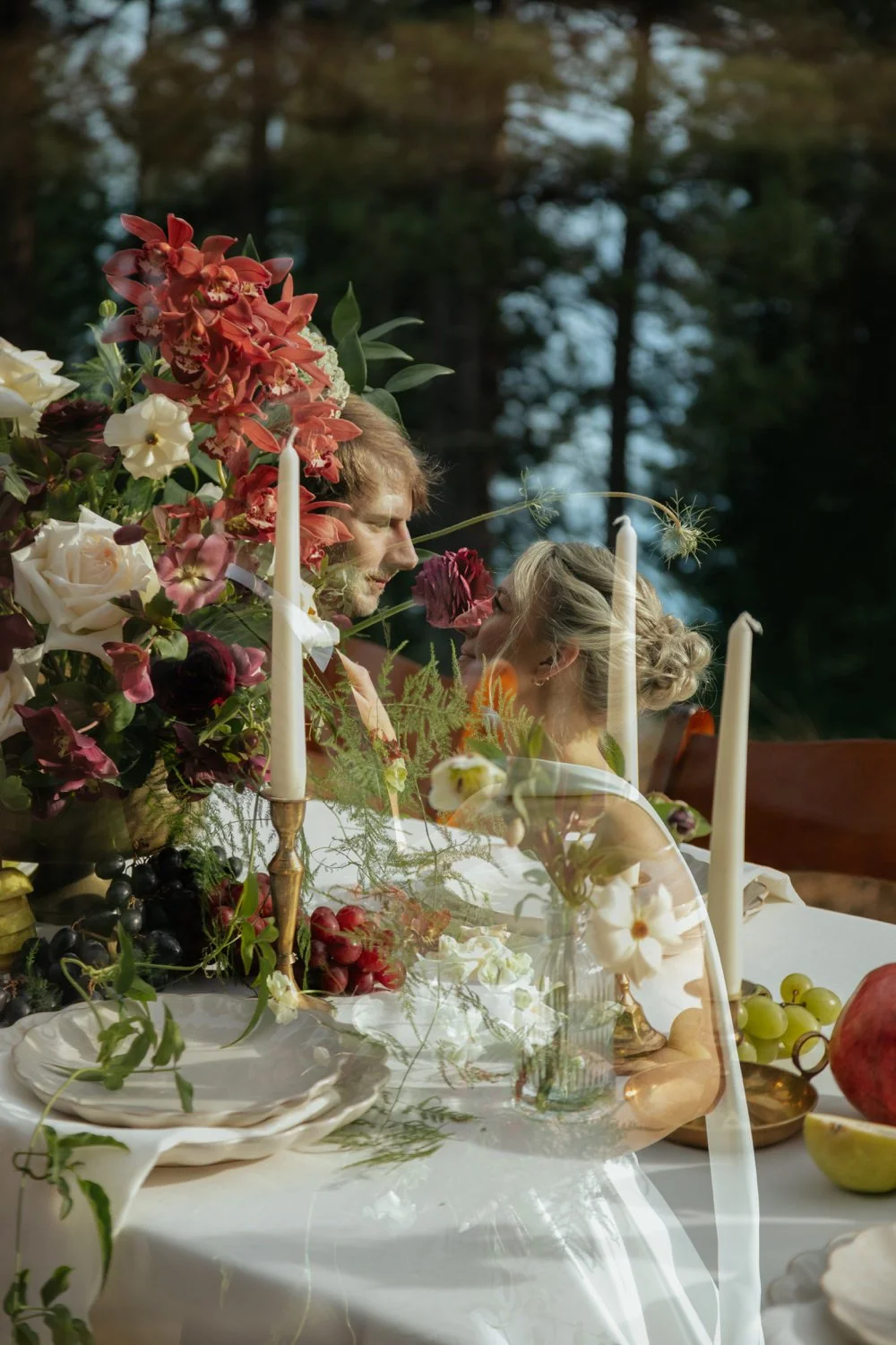 A romantic scene of a couple at a dining table with floral arrangements, candles, and fruit outdoors, with trees in the background.