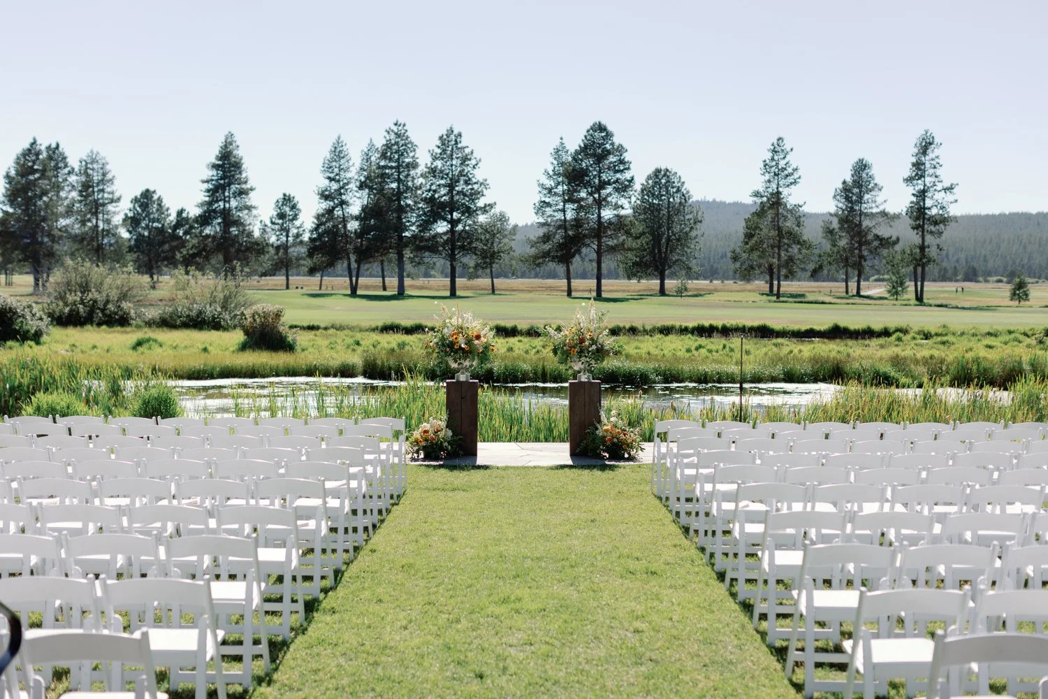 Outdoor wedding setup with white chairs arranged on grass facing a pond, with flower arrangements on wooden pedestals, overlooking a field and trees in the background.