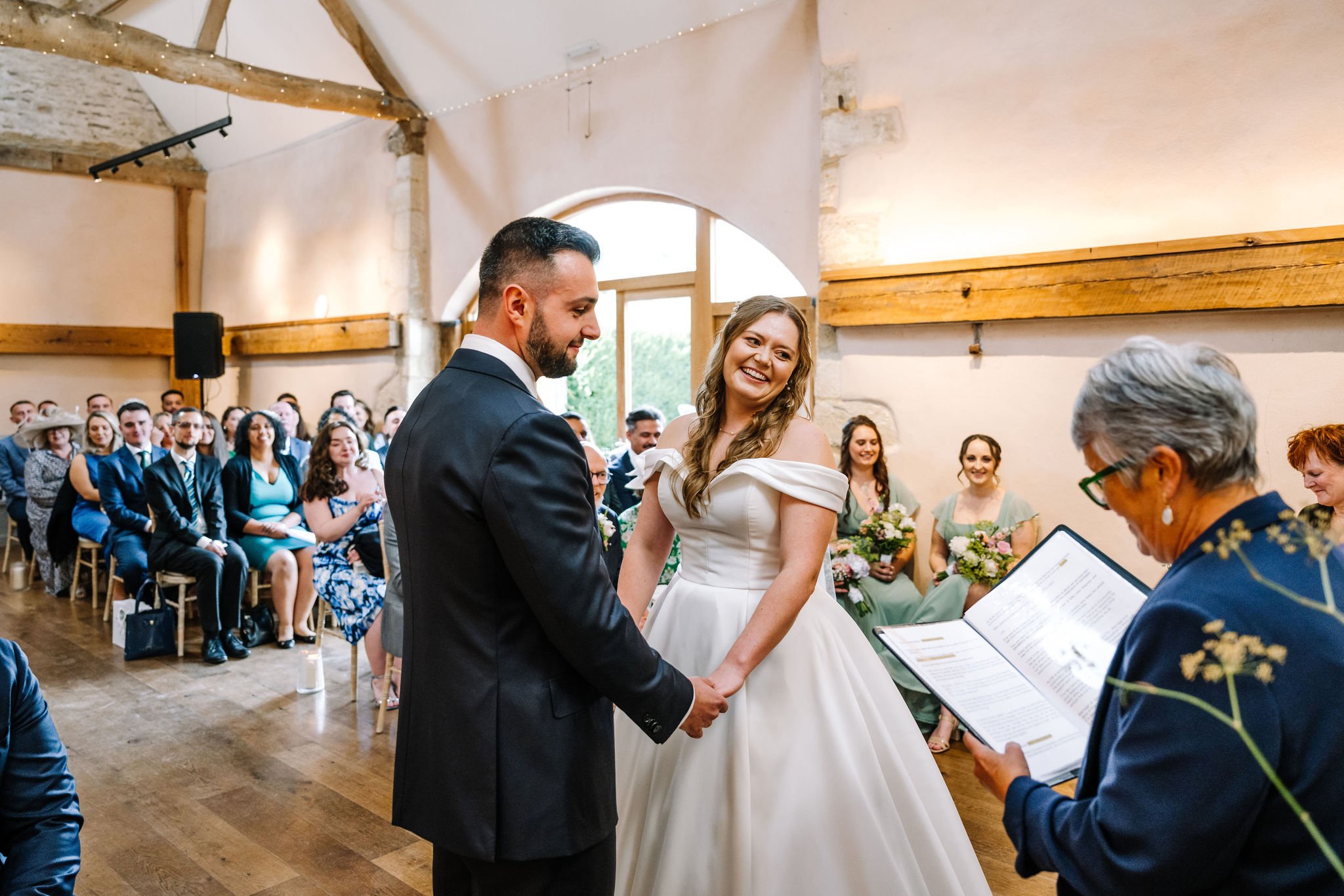 A wedding ceremony with a bride and groom holding hands, smiling at each other. The ceremony is taking place indoors with guests seated in the background. A person is reading from a booklet, likely a minister or officiant. The setting is a rustic venue with wooden beams and soft lighting.