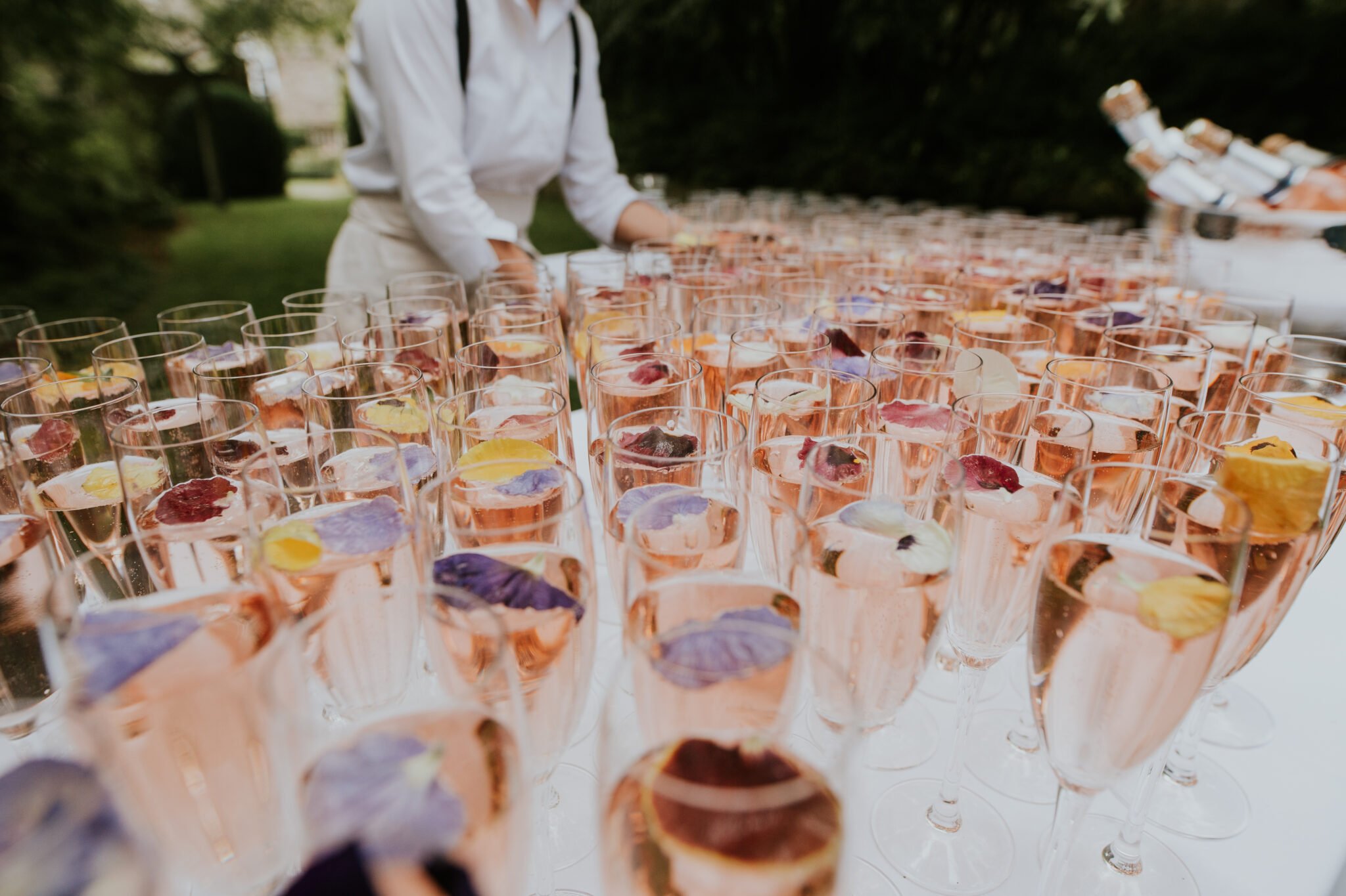 Rows of champagne glasses with pink liquid and floating flower petals, served outdoors, with a person in a white shirt arranging them.