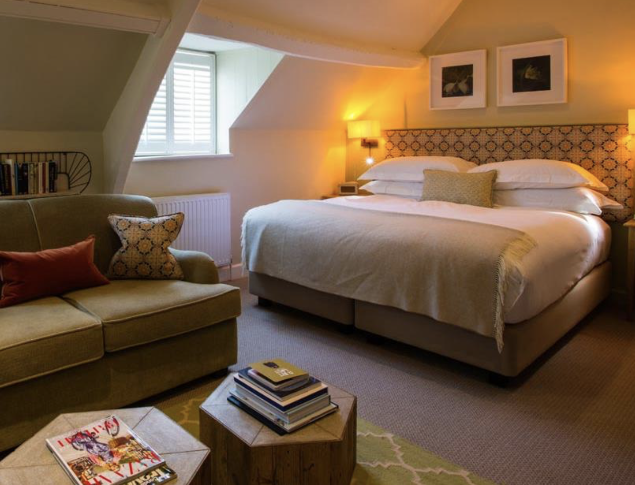 Cozy bedroom with a double bed, pillows, blanket, two wall lamps, couch, books, window, and a small table with magazines.