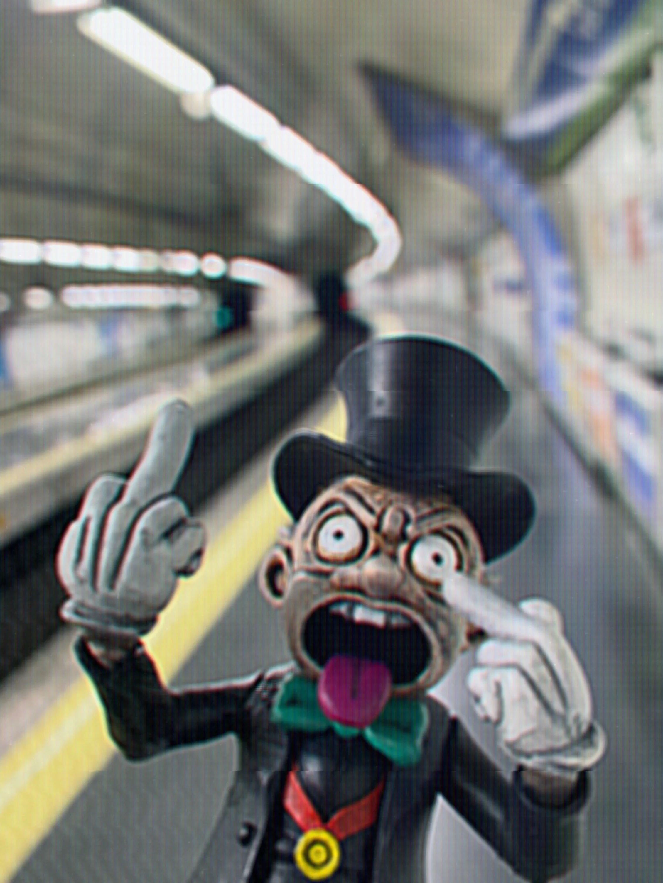 A person dressed in a clown costume with a large head, exaggerated facial features, and a green bow tie, standing on a museum or store aisle, making a shock gesture with both hands, with shelves and lighting above.