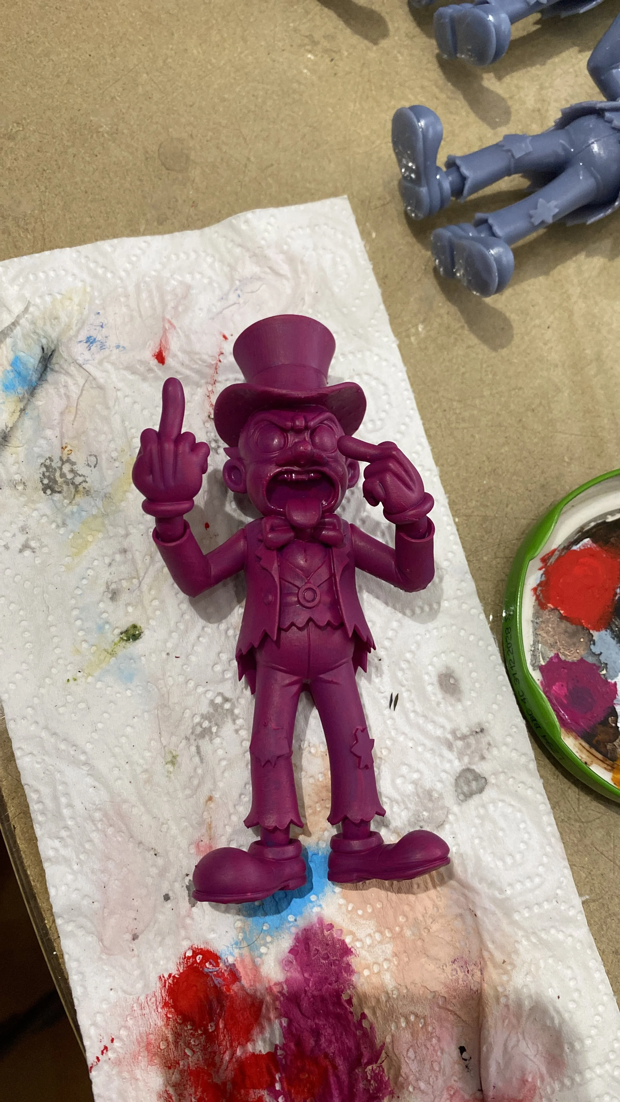A purple figurine of a clown with exaggerated features, wearing a top hat, bow tie, and patched clothes, making a rude gesture with one hand and pointing to its eye with the other, lying on a paper towel with paint stains.