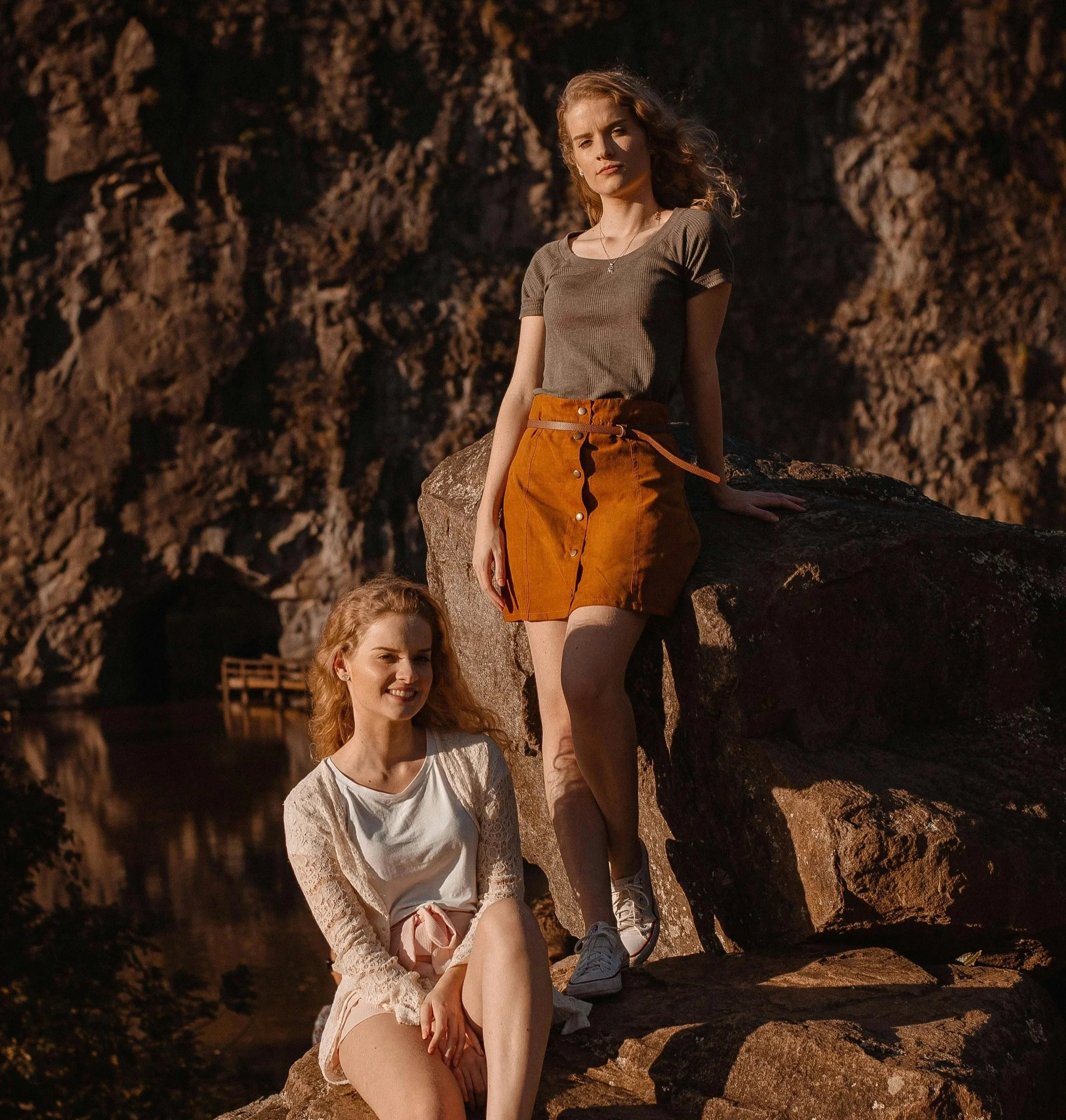 Young women sitting and standing on rocks by a river at sunset, enjoying outdoor scenery.