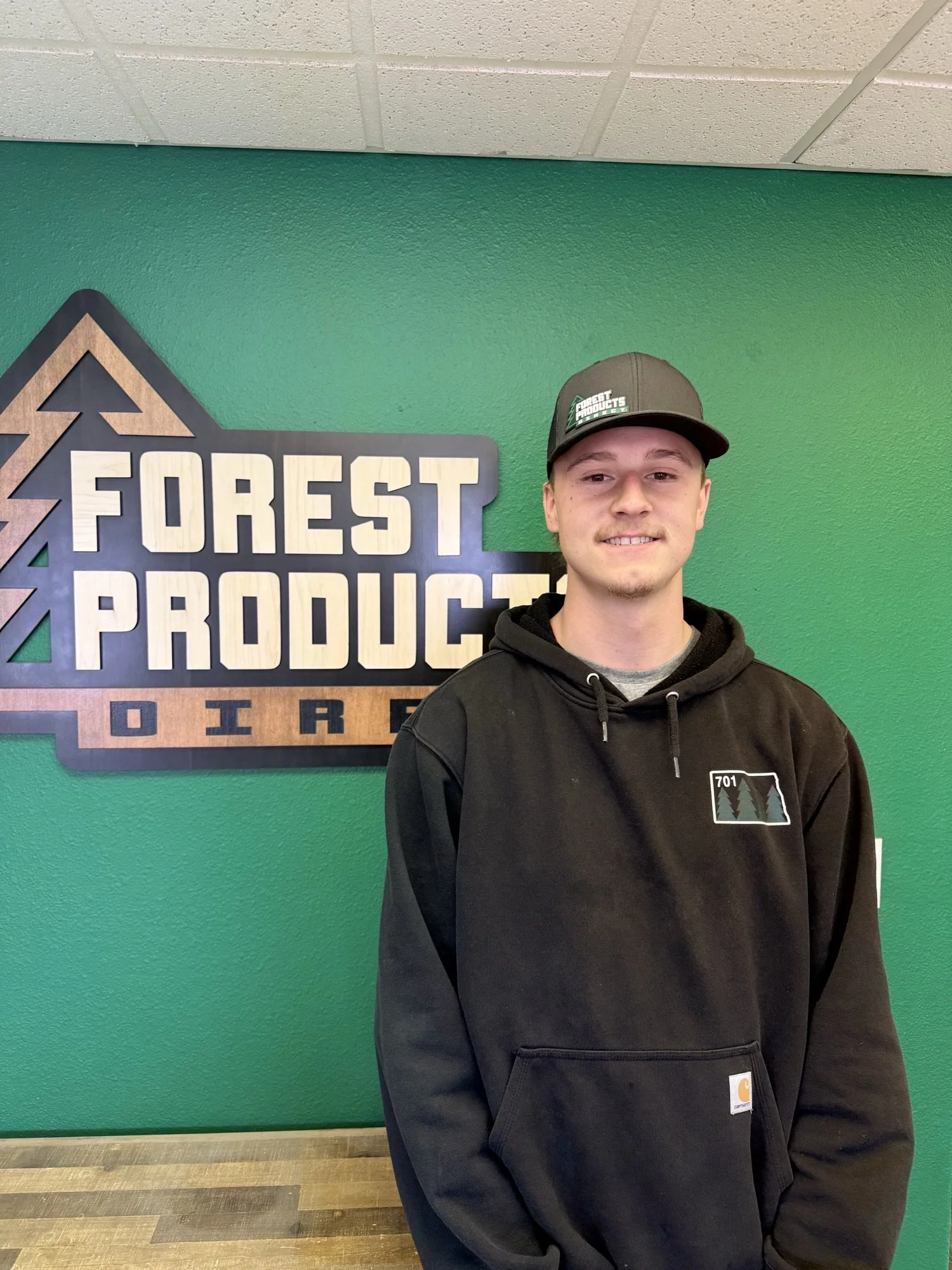 A young man standing in front of a green wall with a sign that says 'Forest Products' and a tree logo. He is wearing a black hoodie with a mountain and tree design on the front, and a black baseball cap with a green and white logo that reads 'Forest Products'.