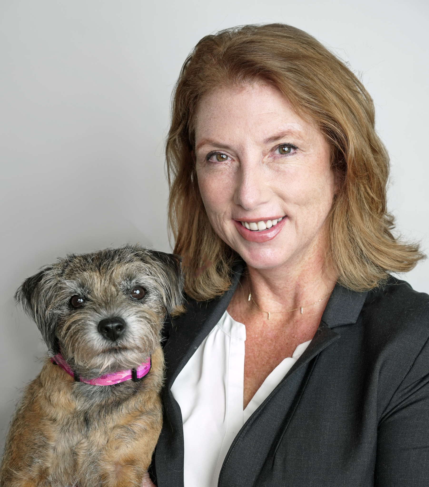 A smiling woman with light skin, reddish-brown hair, and brown eyes, wearing a black blazer and white blouse, holding a small furry dog with a pink harness against a light styled background.
