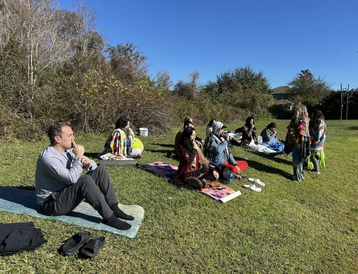 A group of people sitting on grass outdoors, practicing breathing exercises and meditation. Some are sitting on mats and blankets, others are wrapped in colorful blankets. The background features trees and clear blue sky.