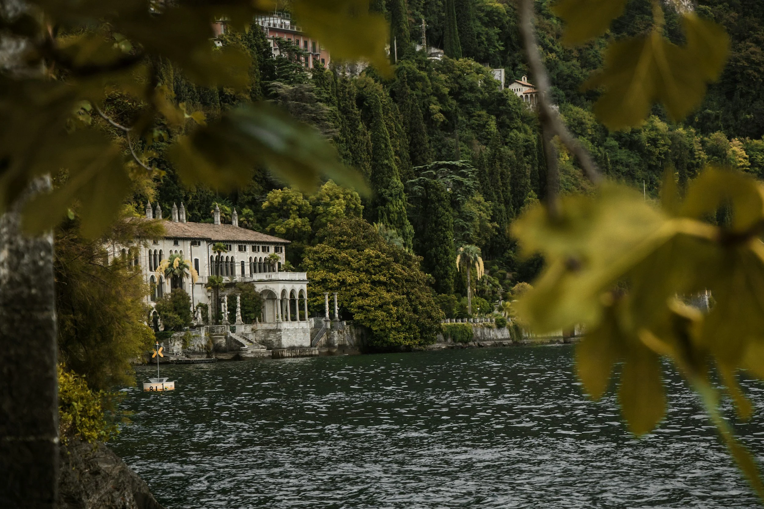 A lakeside estate surrounded by lush greenery and tall trees, with a large white villa featuring arched windows and balconies, partially obscured by blurred leaves in the foreground.