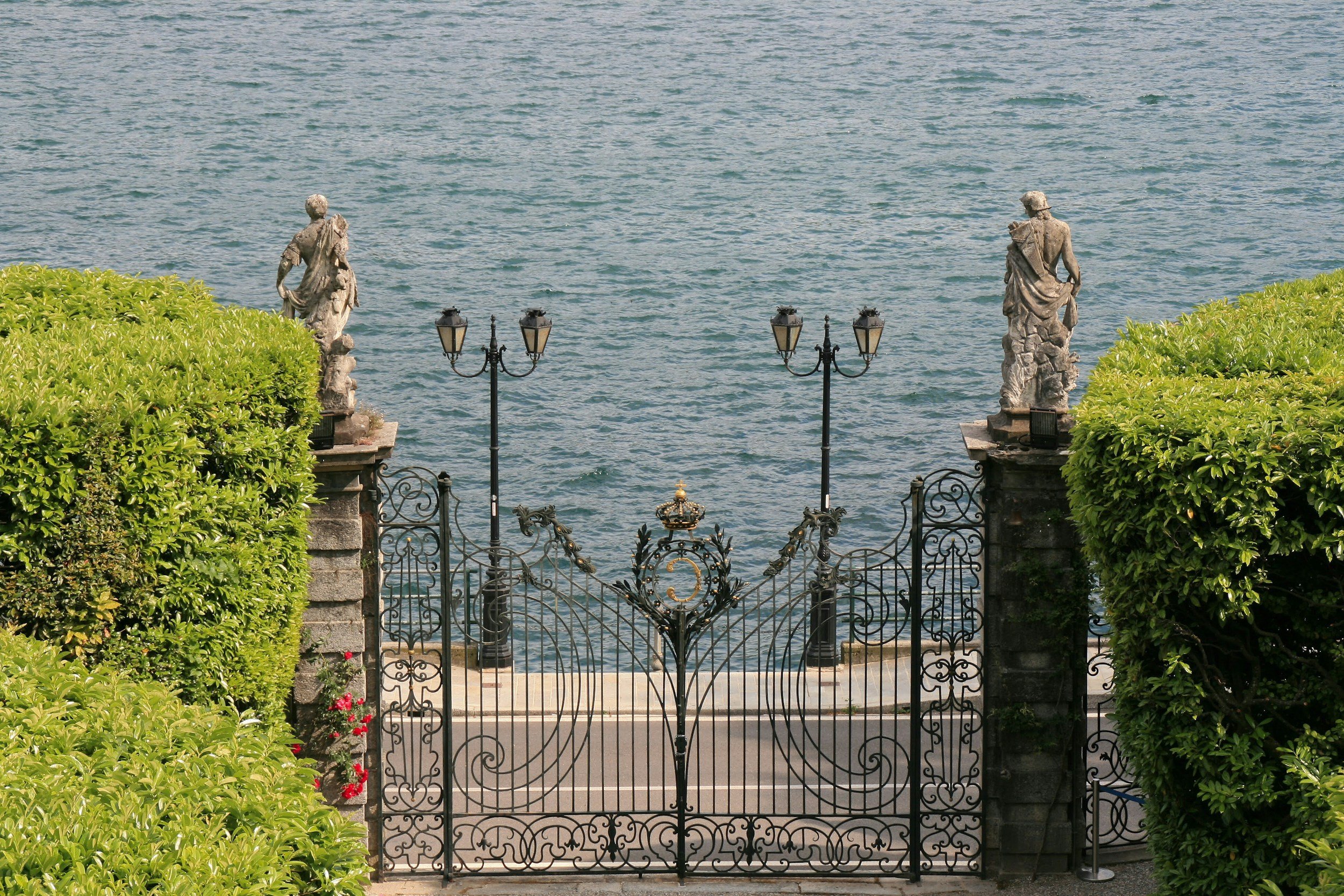 Decorative wrought iron gate with ornate details, flanked by two stone statues on pedestals, opening to a water view
