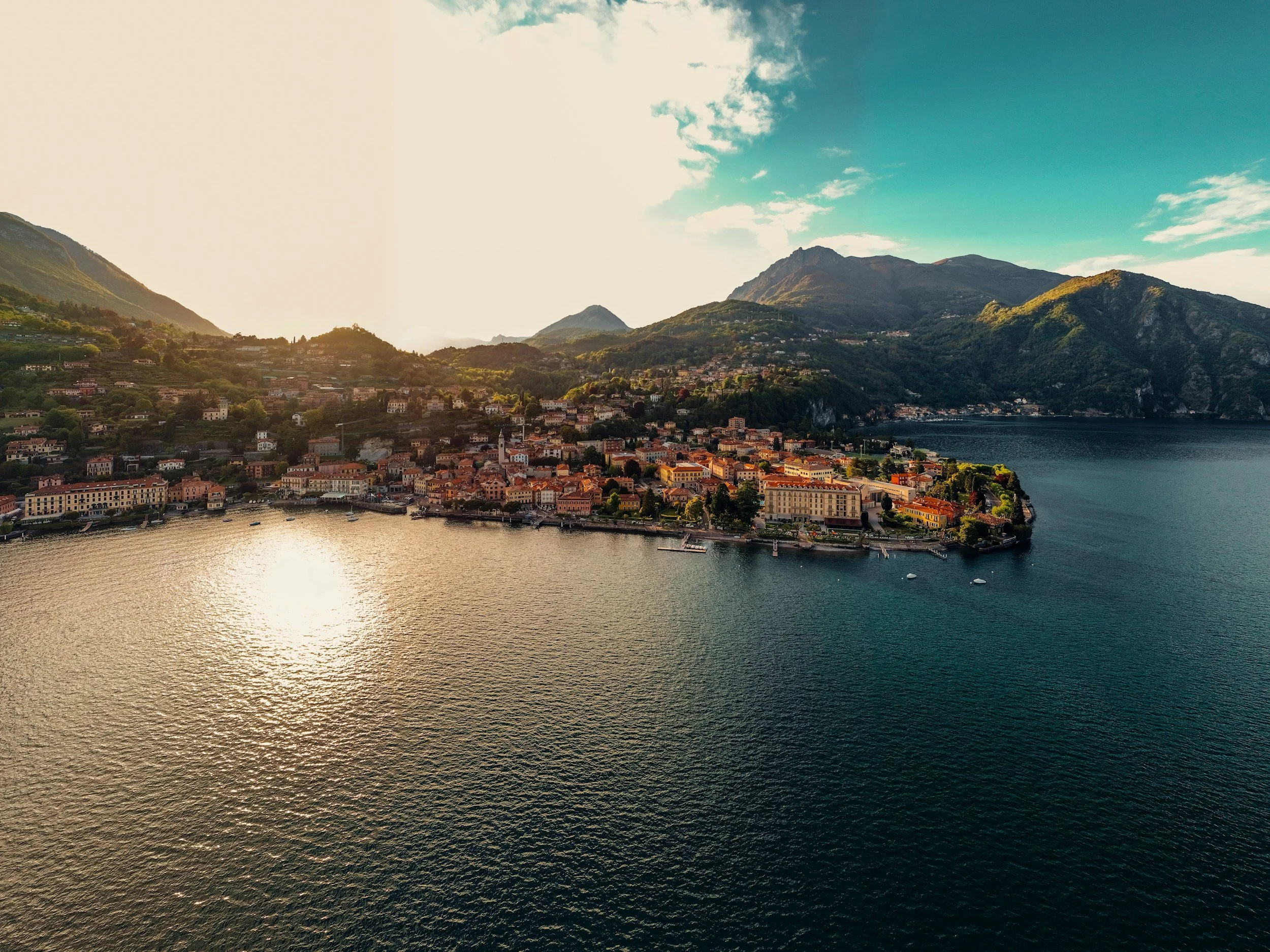 Aerial view of a lakeside European town with colorful buildings, surrounded by green mountains and a partly cloudy sky.