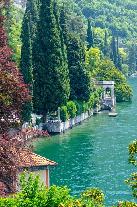 Lush greenery along a lakeside with trees, a stone wall, and a decorative structure by the water.