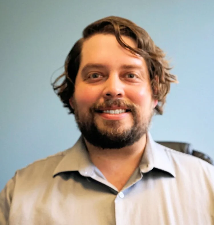 A smiling man with wavy brown hair and a beard, wearing a light gray collared shirt, in front of a light blue wall.