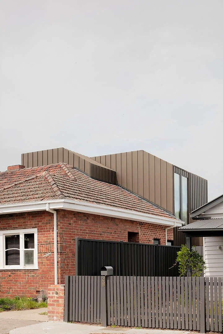 Contemporary metal-clad upper-level addition rising behind a red brick heritage home, contrasting old tiled roof with modern vertical cladding