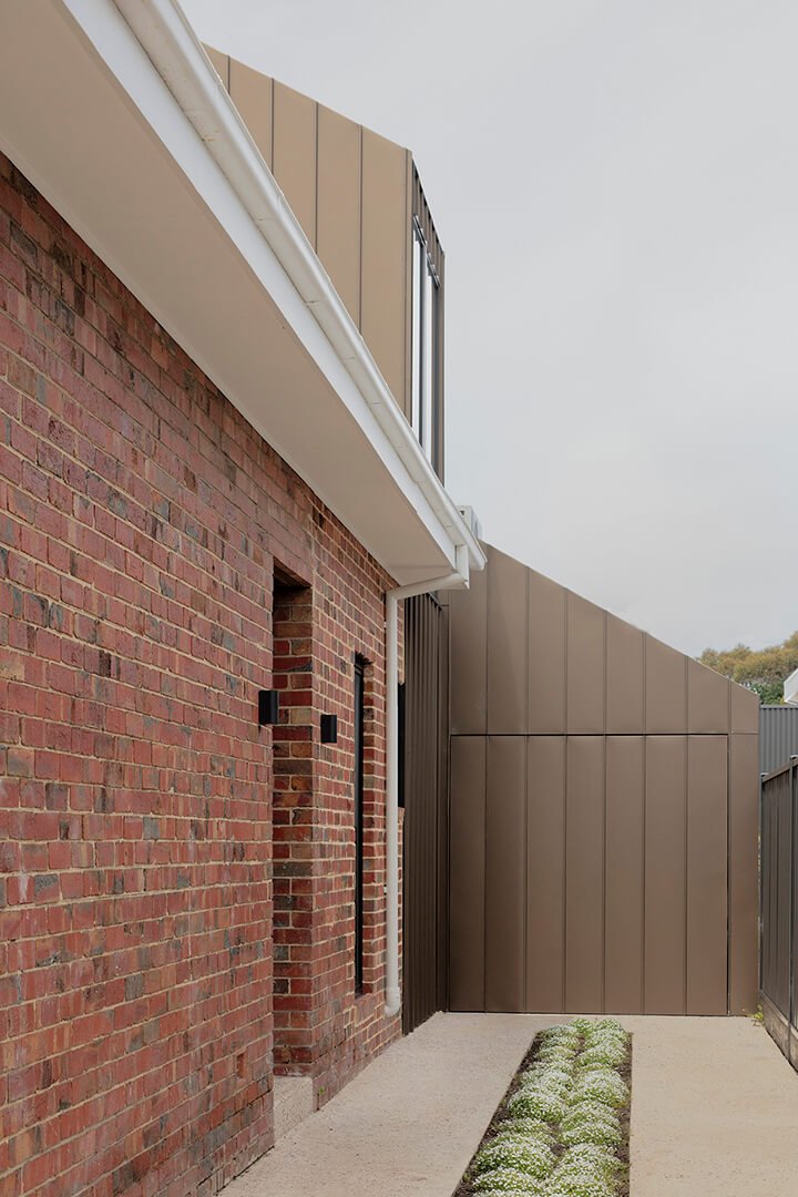 Street view of renovated brick house with new sculptural upper volume in bronze-toned metal cladding set behind the original roofline and front fence
