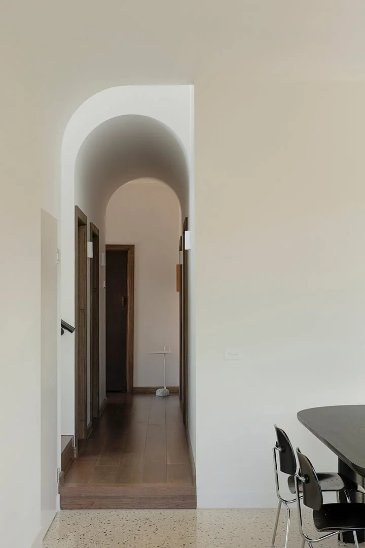 Minimalist interior with a white arched hallway leading to the original part of the home. Foreground shows a contemporary dining area with black chairs and a table.