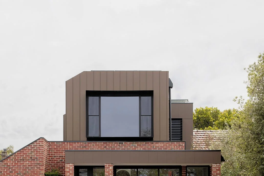 Modern house exterior with a geometric design featuring a bronze metal upper structure, large central window, and red brick lower facade, set against a cloudy sky.