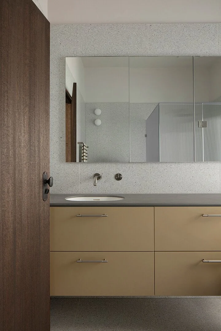 Modern bathroom with a minimalist design featuring a large mirror, beige vanity with sleek handles, gray countertop, and stainless steel tap; wood door partially open.