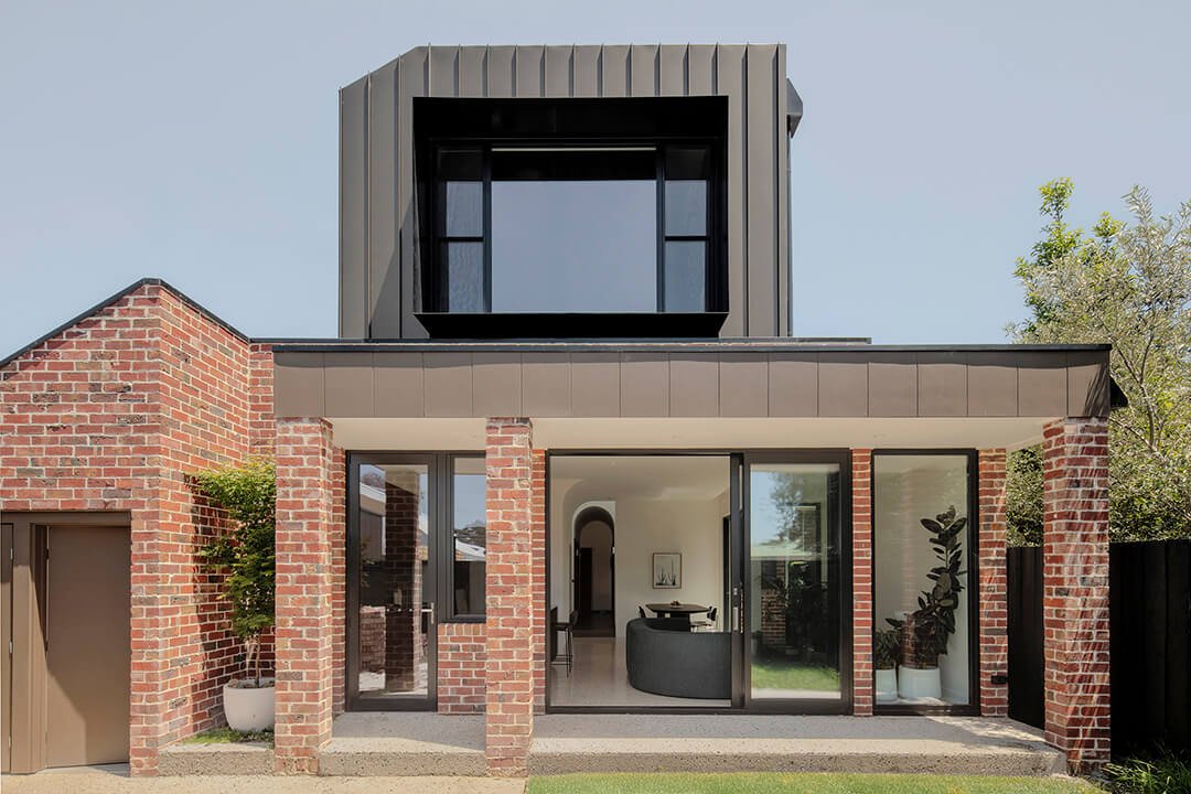 A modern two-story house featuring a mix of red brick and dark cladding. Large windows and sliding glass doors create a light, open atmosphere.